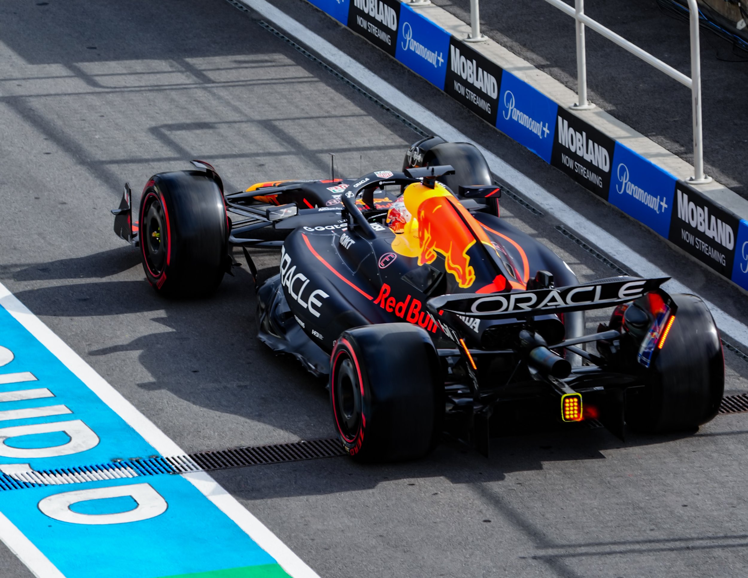 A Formula 1 race car with Red Bull Racing logo and Oracle sponsorship is at the pit stop, next to a blue and black advertisement sign for Movbland and Paramount+ streaming service, on a race track with white and blue markings.