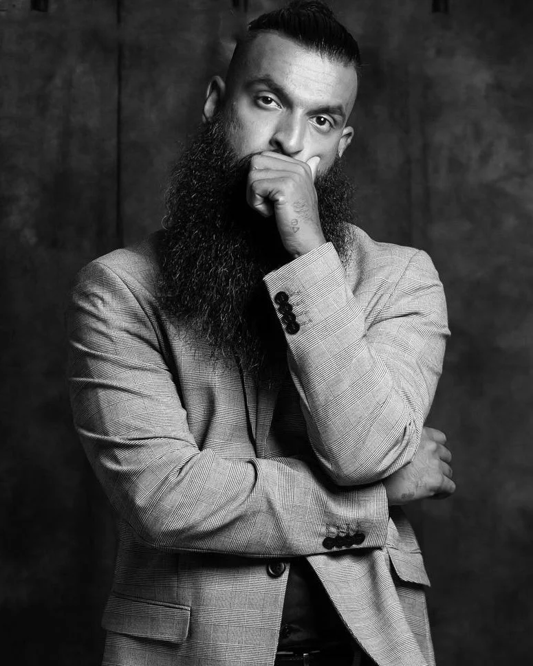 Black and white portrait of a man with a beard, wearing a checked blazer, with his hand touching his chin, looking thoughtful.