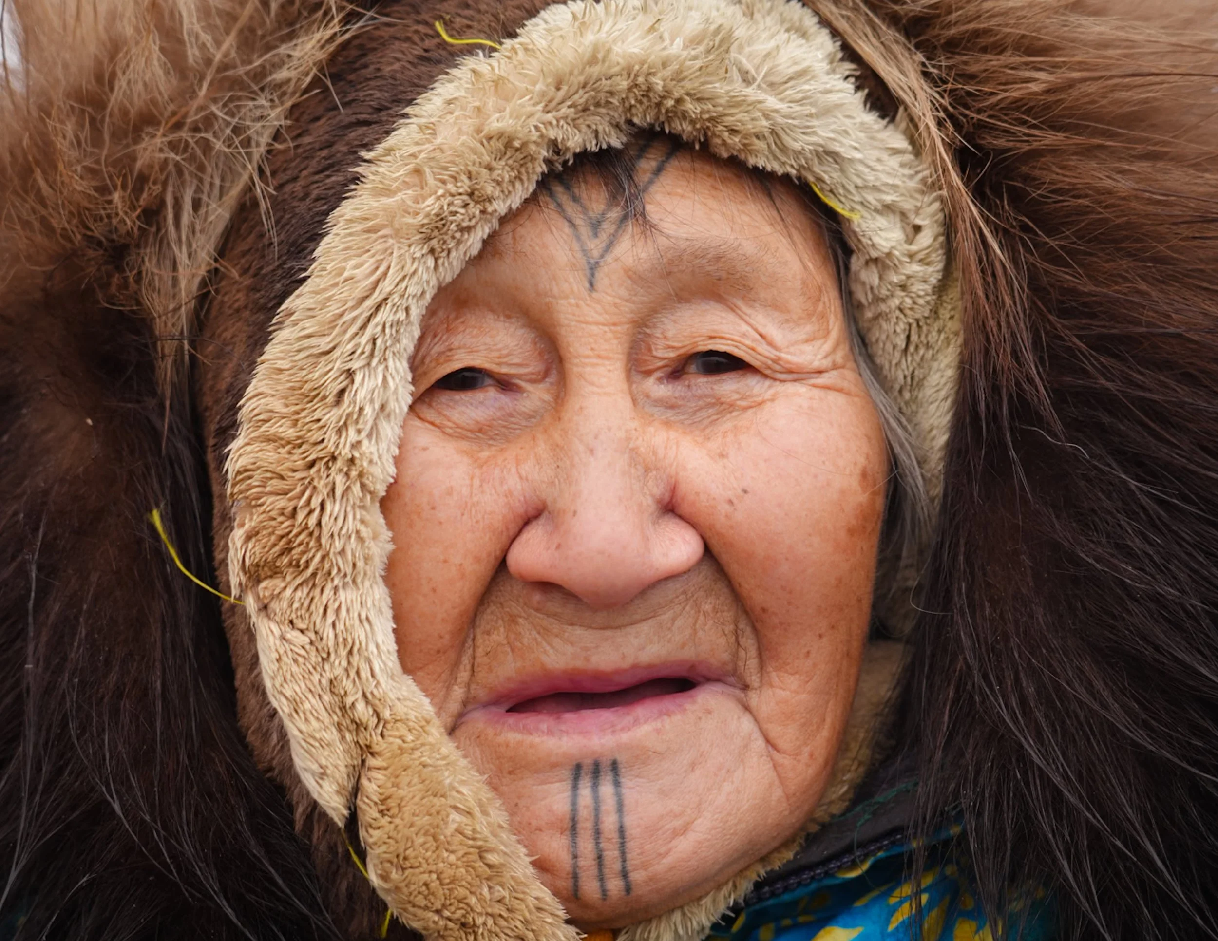 Close-up of an elderly woman wearing a warm, furry hooded coat with a pattern of brown and tan fur around her face. The woman has wrinkles and traditional tattoos on her face with vertical lines on her chin. The background is blurry, likely outdoors.