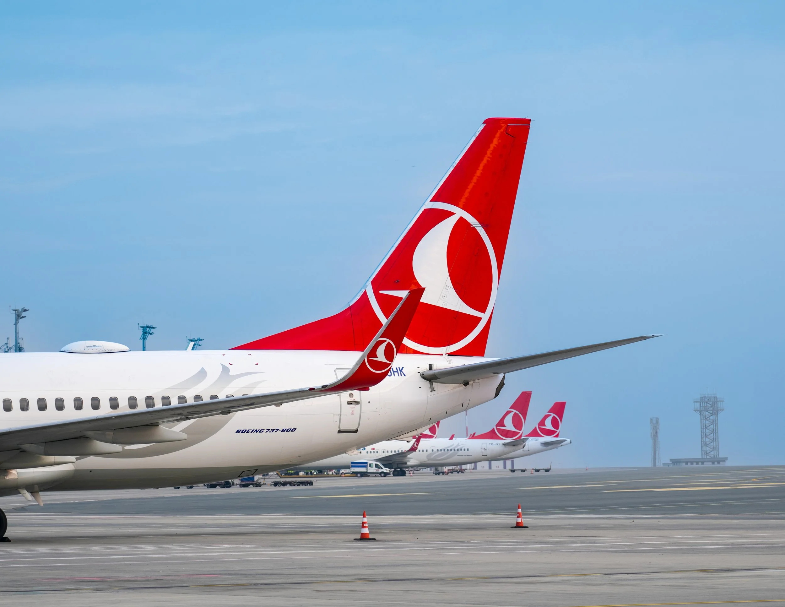 Multiple airplanes with red tail fins parked at an airport runway, with orange traffic cones on the ground and some airport structures and equipment in the background.