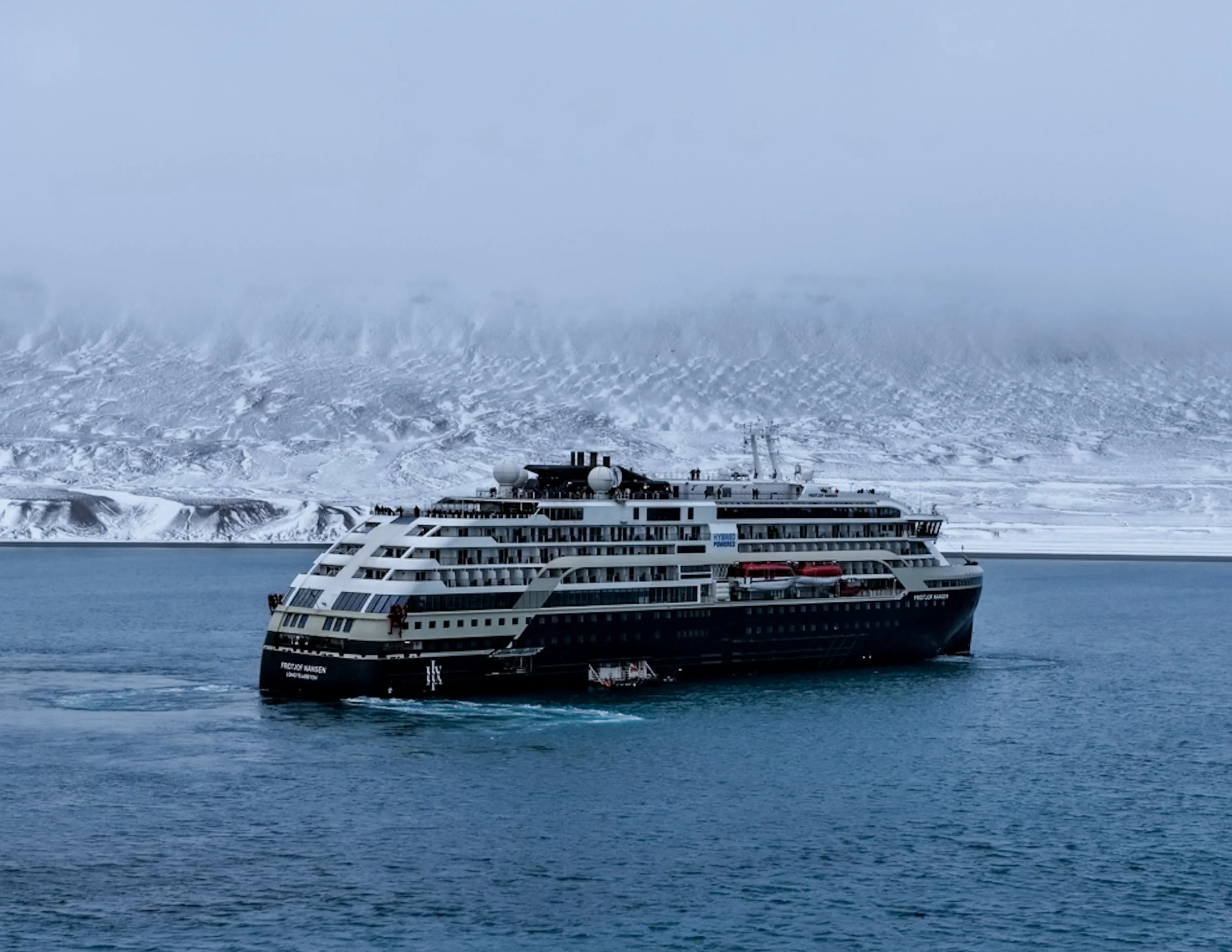 A large cruise ship sailing in icy waters with snow-covered mountains in the background and a cloudy sky.