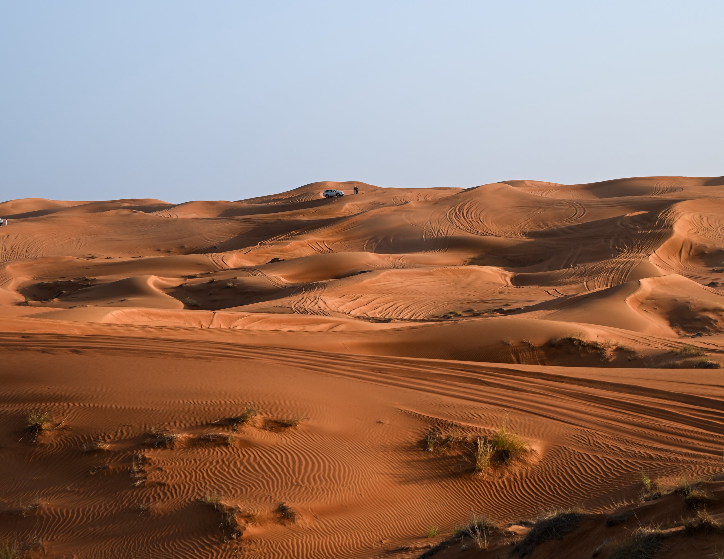 Sand dunes in a desert with a vehicle and two people standing on a dune, and tire tracks on the sand.
