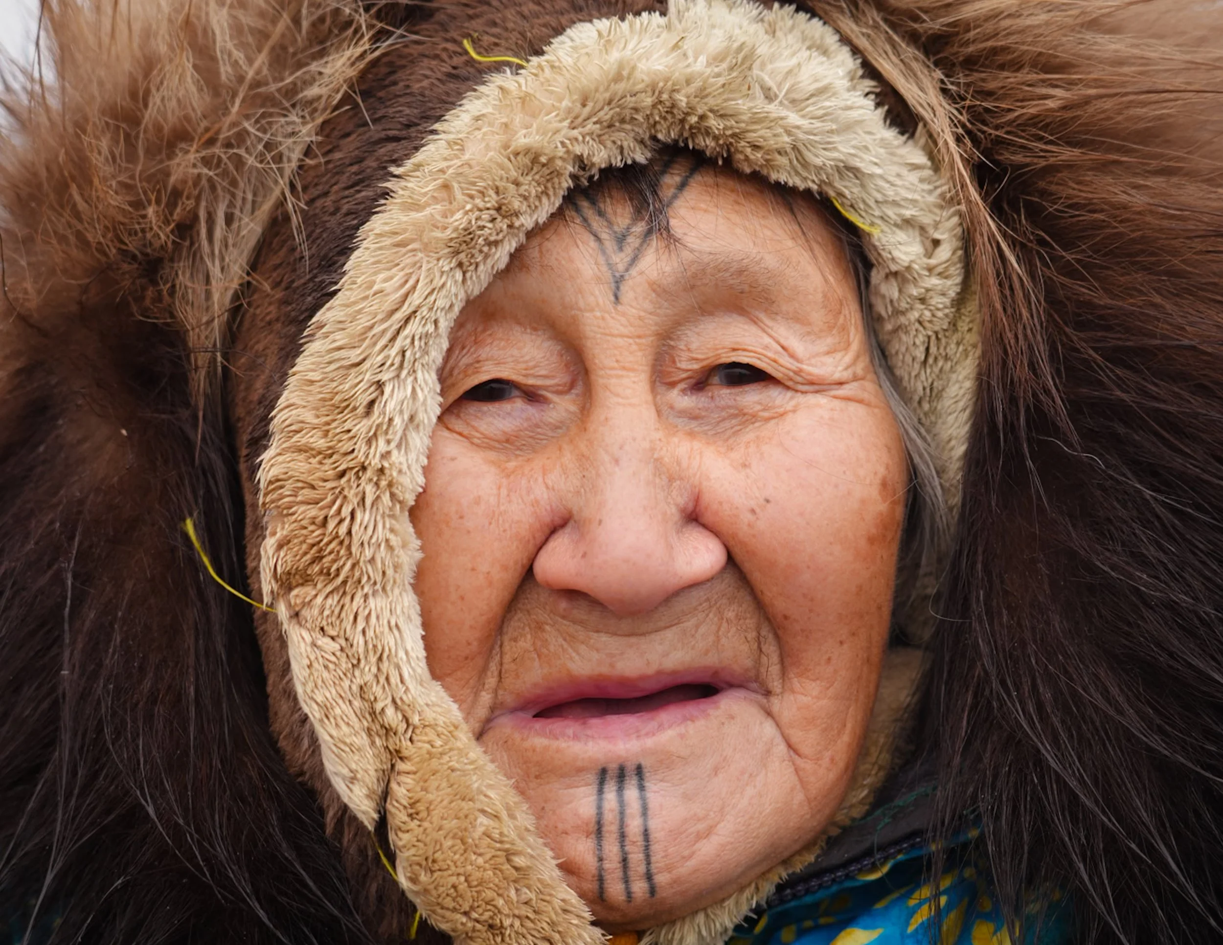 Close-up of an elderly woman wearing a thick, furry hooded coat with leather trim, fur-lined with dark brown and beige colors, and tribal face markings.