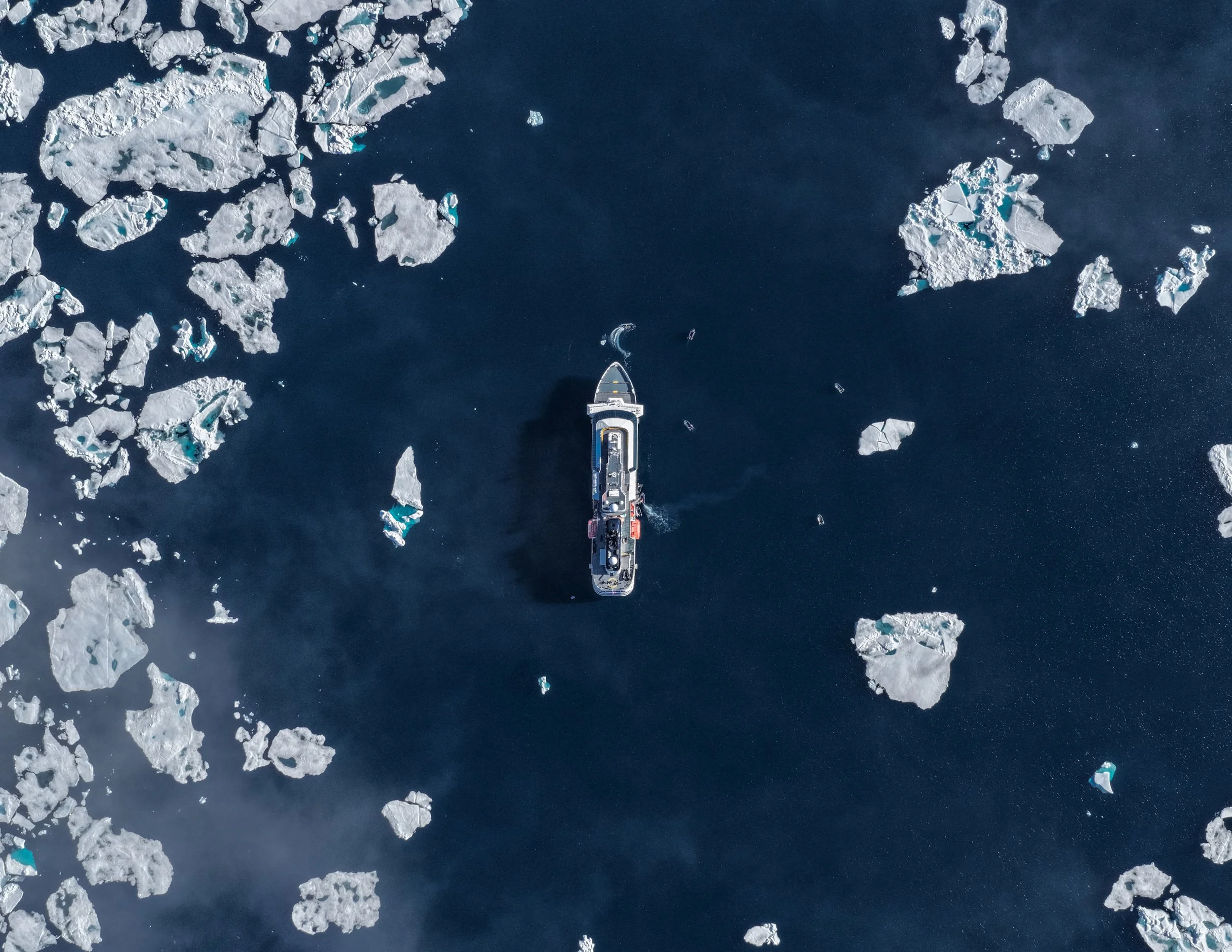 An aerial view of a ship navigating through icy waters surrounded by large floating ice sheets and broken ice chunks.