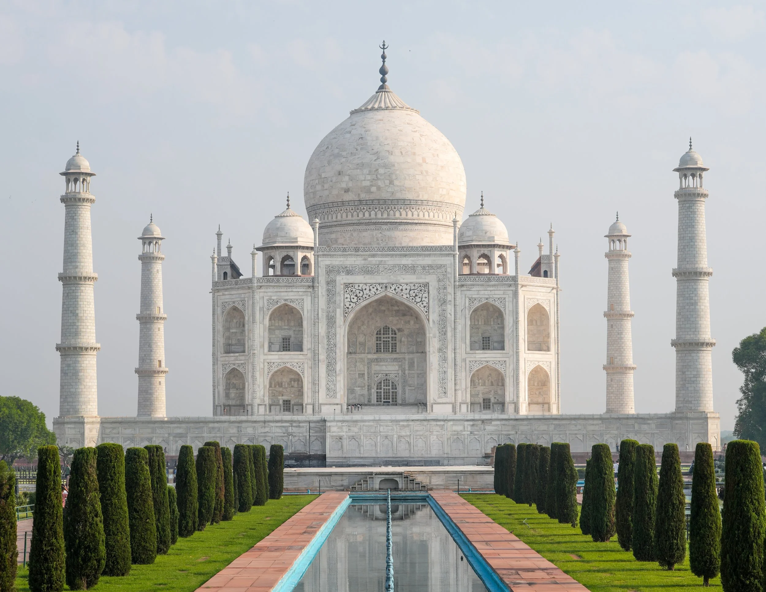 The Taj Mahal, a white marble mausoleum with a large central dome, four minarets at each corner, and intricate architectural details, situated in a garden with a reflecting pool in front.