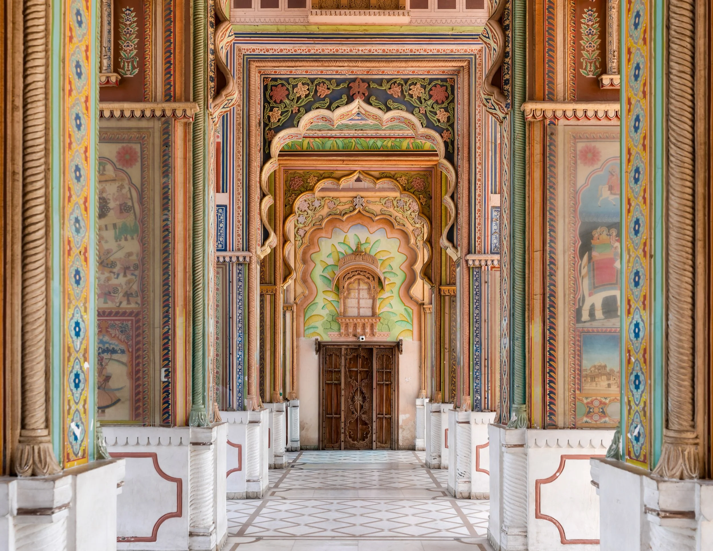 Colorful, ornate corridor with floral and geometric patterns, arches, and decorative columns in a traditional Indian architectural style.