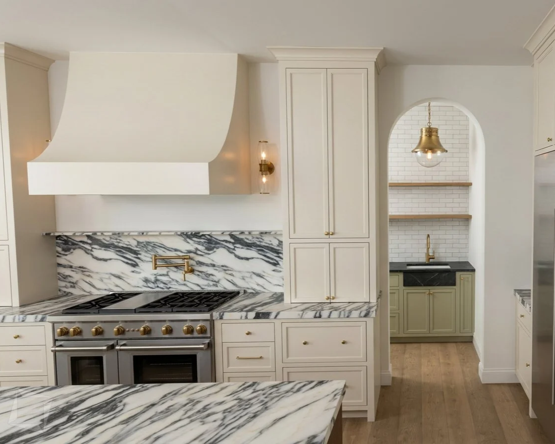 Modern kitchen with white cabinets, marble countertops and backsplash, gold hardware and fixtures, and a small nook area with a black sink and gold faucet.