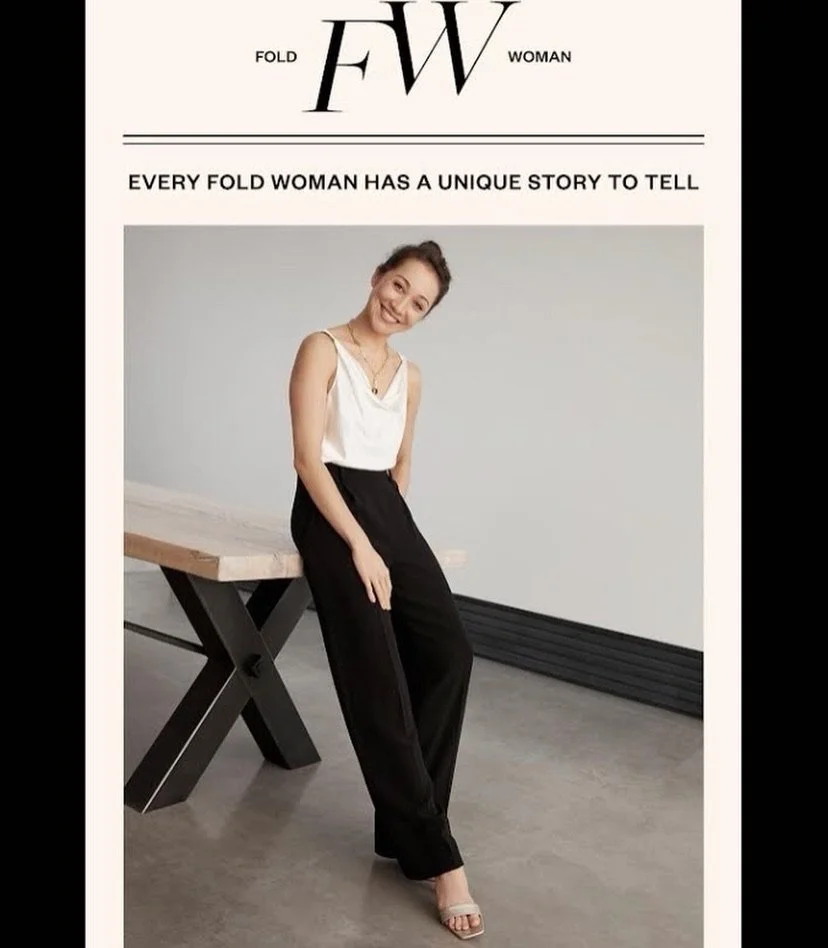 Merritt Moore in a white top and black pants leaning against a table, smiling at the camera in a minimalist indoor setting for Fold women feature.