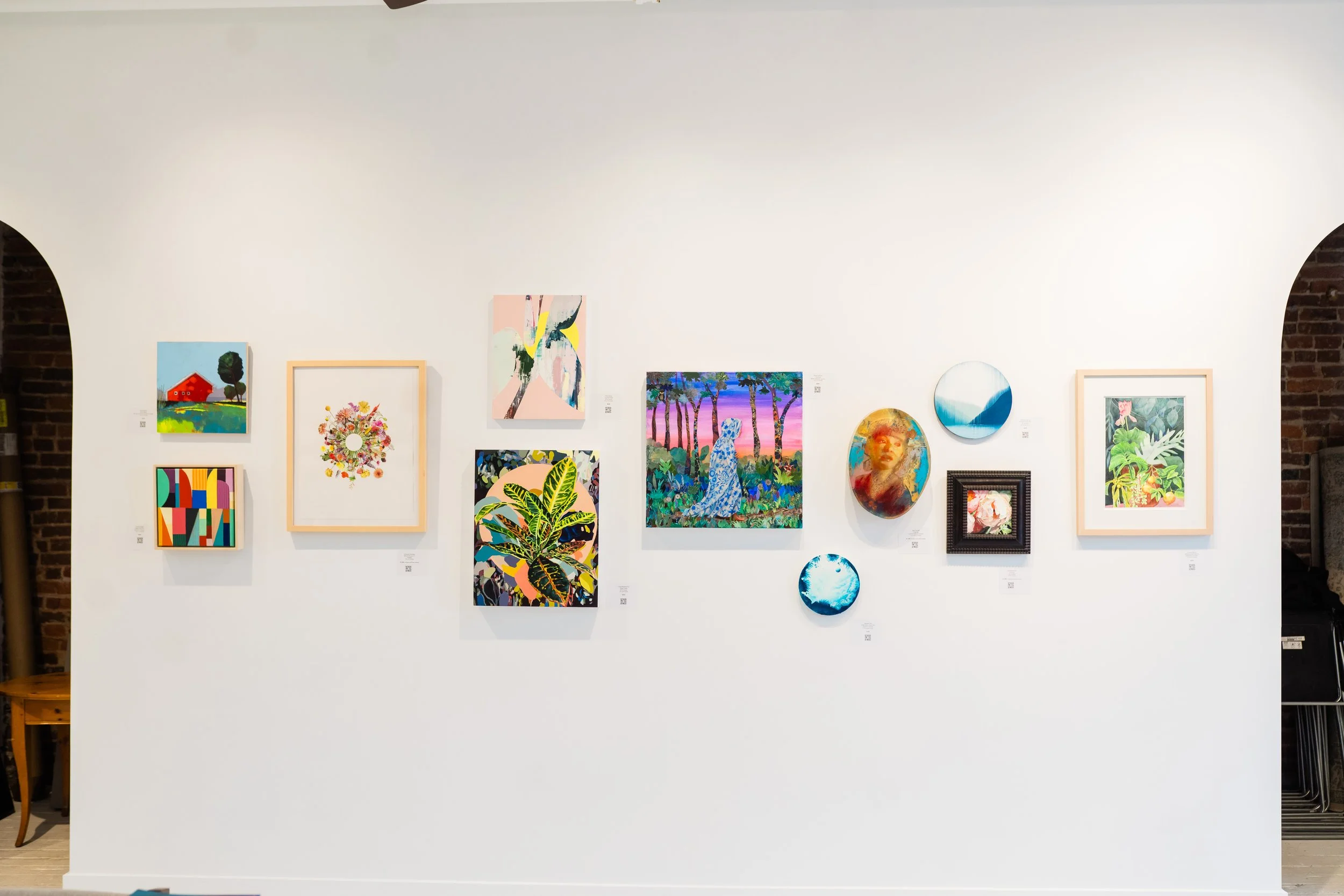Gallery wall with various colorful paintings and circular artworks displayed on a white wall in an art gallery.