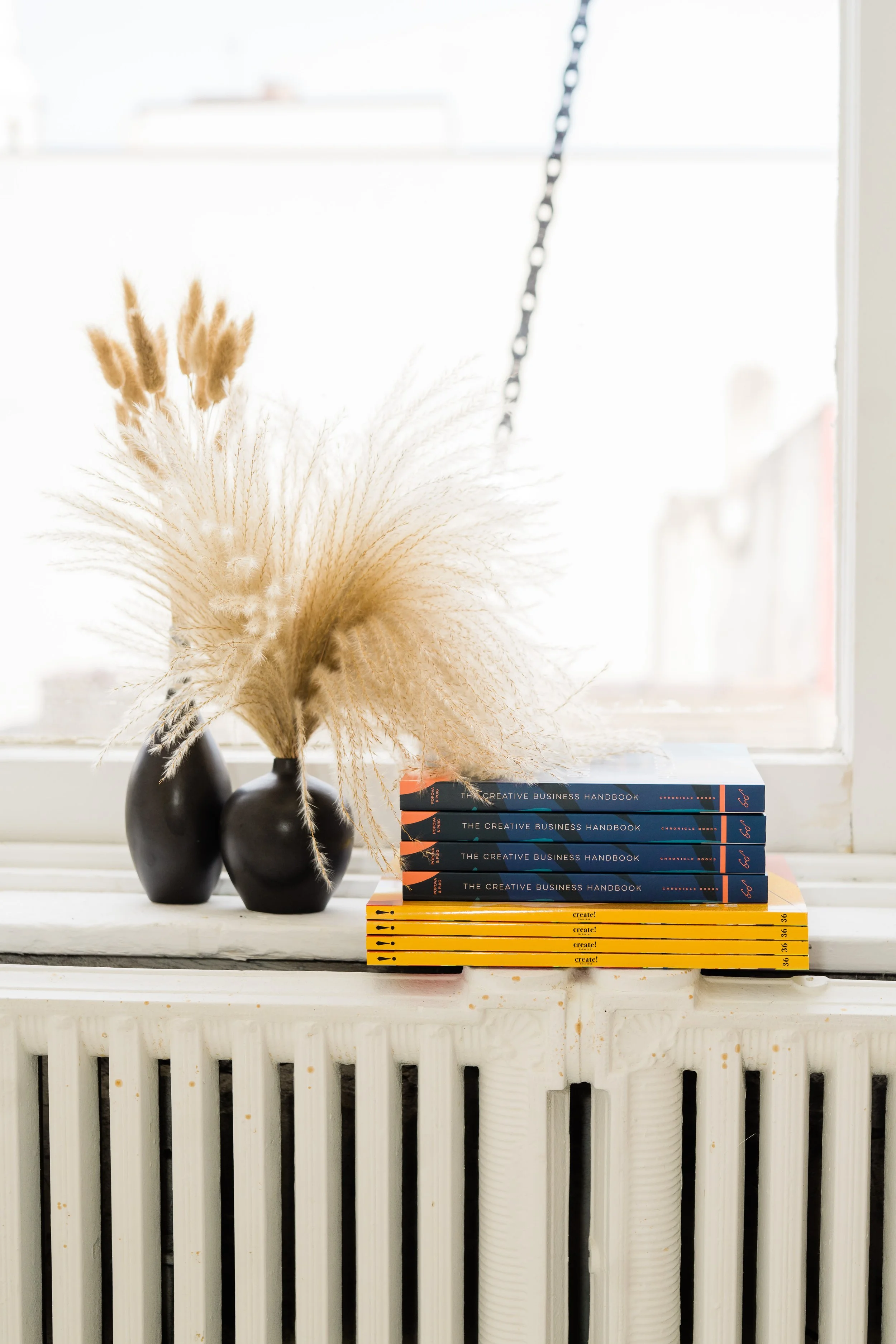 A white windowsill holding three black vases with dried pampas grass, and a stack of ten books titled "The Creative Business Handbook" and "Create!" in yellow.