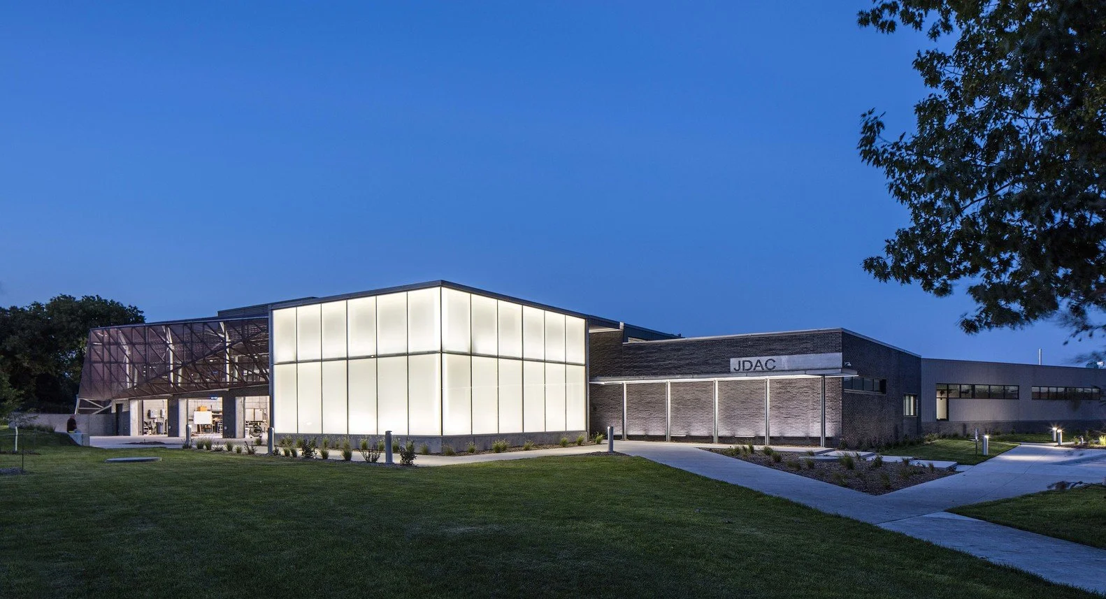 Modern building with illuminated white panels, glass sections, and the letters JDAC on the exterior, surrounded by grass and pathway, under a clear early evening sky.