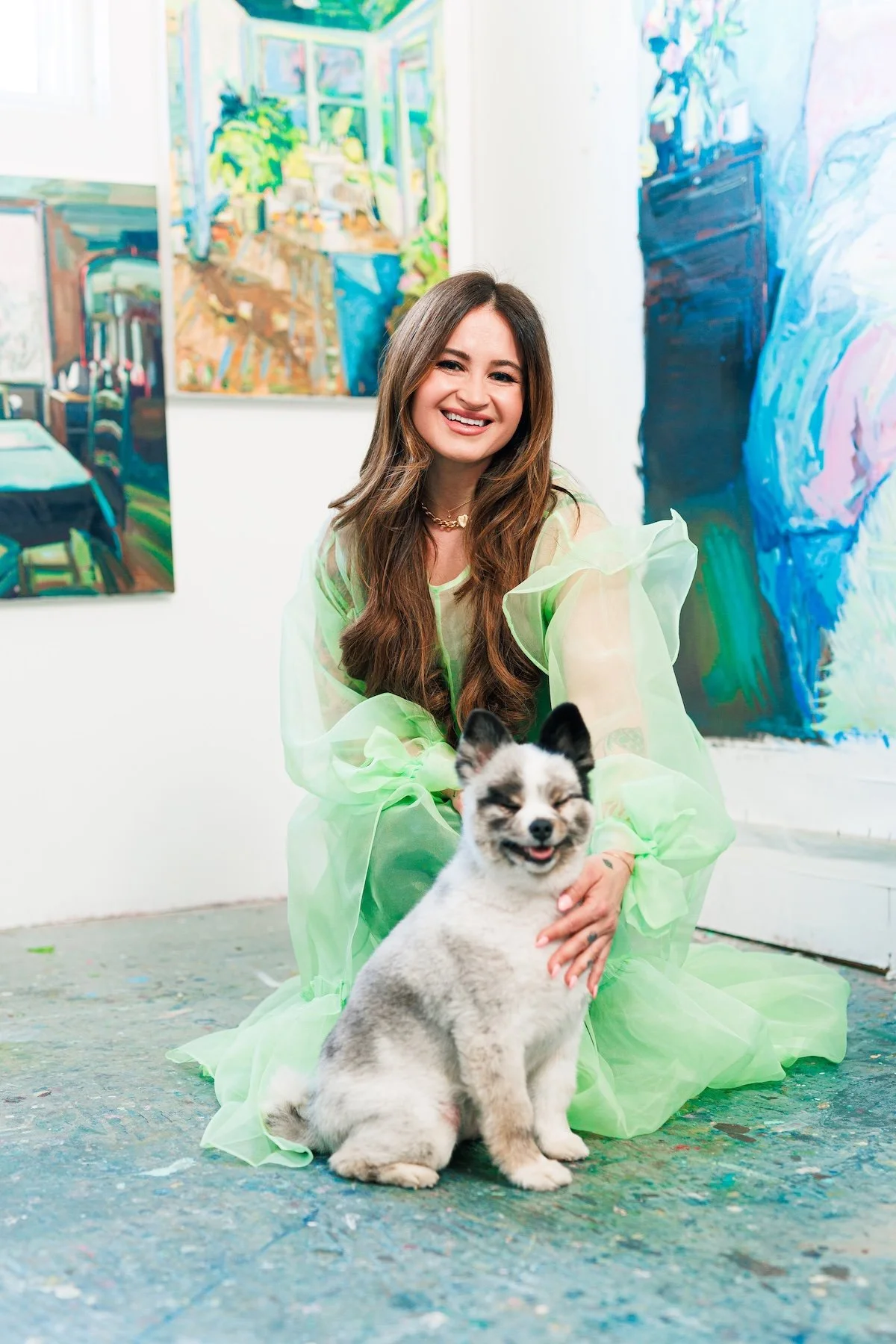 A woman with long brown hair smiling and sitting on the floor in an art gallery, wearing a light green sheer dress. She is holding a cute, smiling puppy with black and white fur, sitting on the floor. The background features colorful abstract paintings.