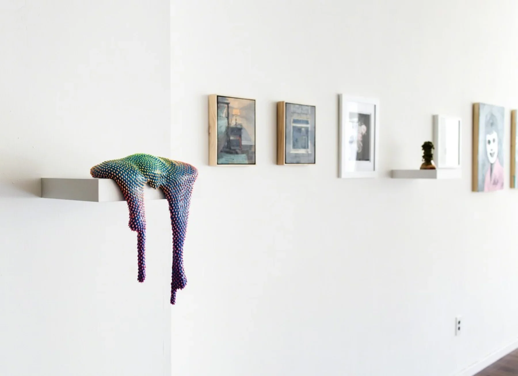 Gallery wall with multiple framed artworks, a small potted plant on a white shelf, and a colorful, dot-patterned textile draped over a side shelf.