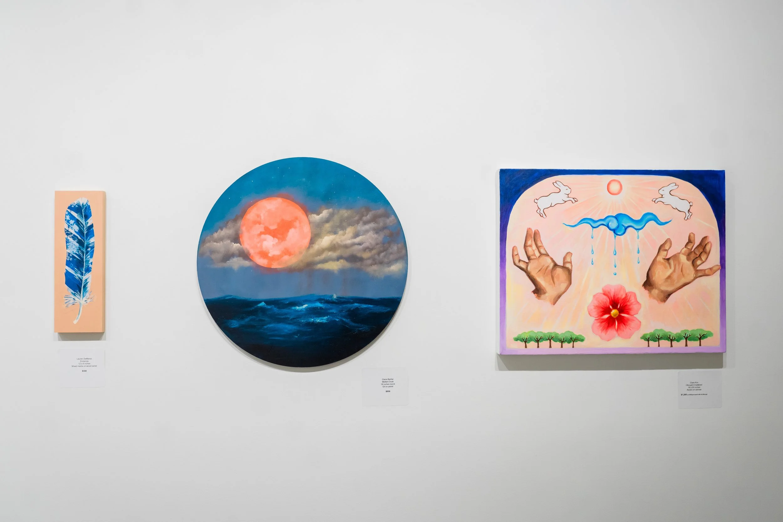 Three paintings on a white gallery wall. The first is a vertical painting of a blue feather on a pink background. The second is a circular painting of a red moon over a dark blue ocean with clouds. The third is a square painting depicting hands, a blue snake-like shape, white rabbits, a red flower, and green trees, with a pink and purple background.