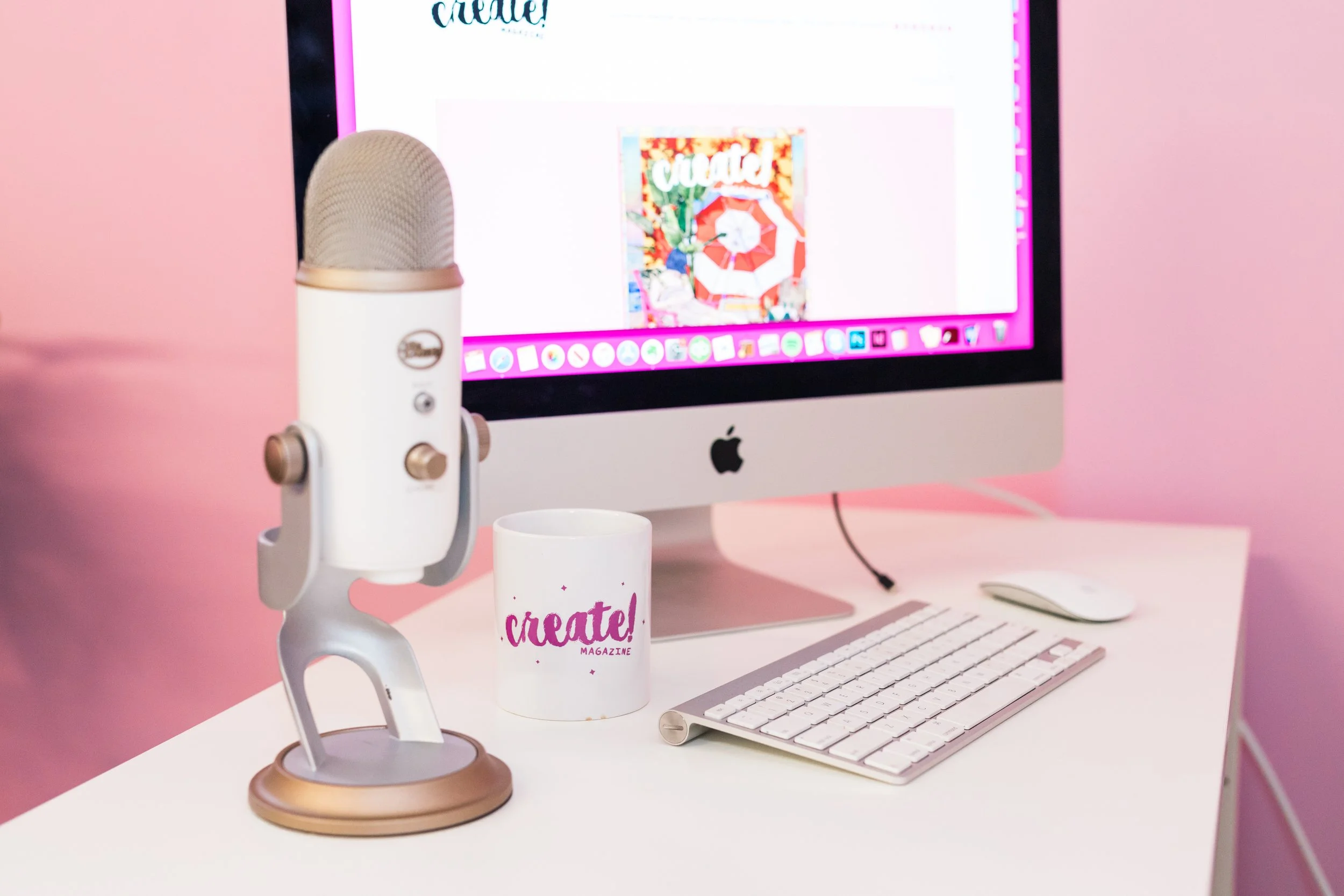 An Apple computer on a pink desk with a white keyboard, a white mouse, a white microphone, and a white mug with pink text that says 'create!'. The computer screen displays a webpage with a colorful cover and the word 'create!' at the top.