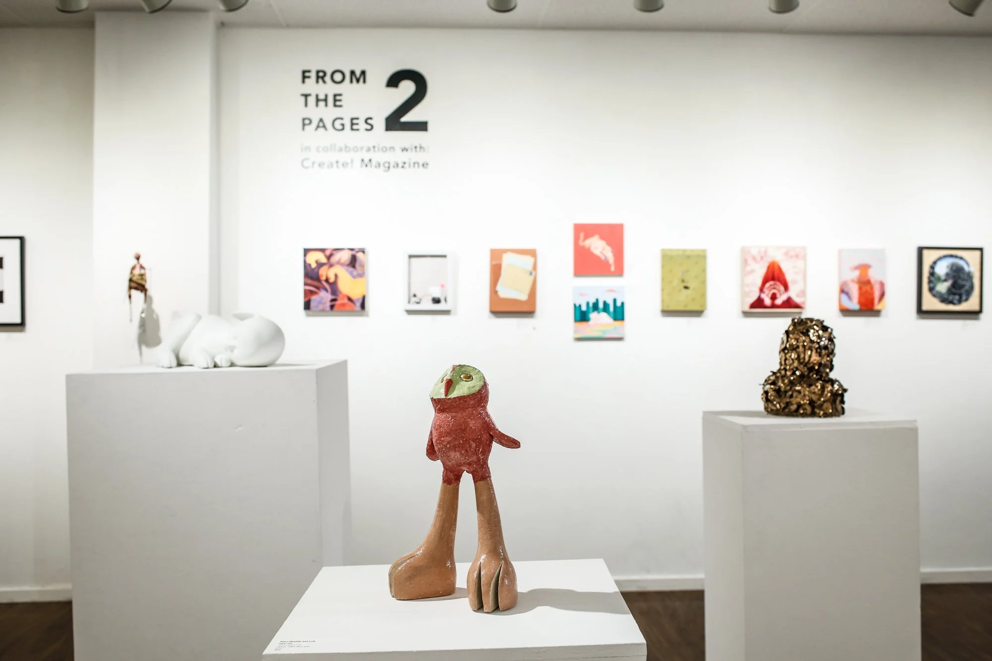 Art gallery with sculptures on pedestals and colorful artwork on the wall, including a ceramic figure with large feet in the foreground.