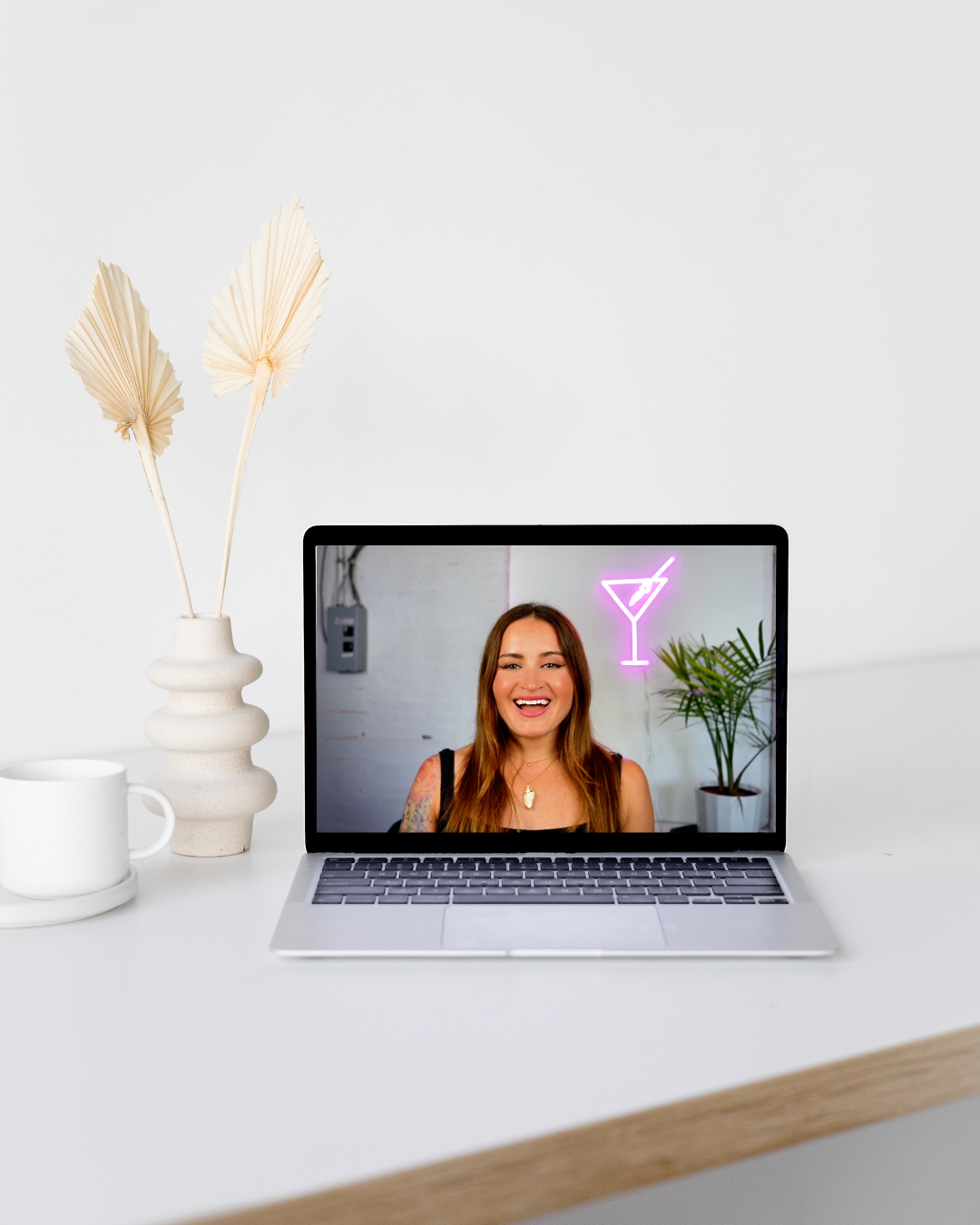 Laptop on a white desk displaying a woman smiling during a video call, with a neon cocktail glass sign and a potted plant in the background.