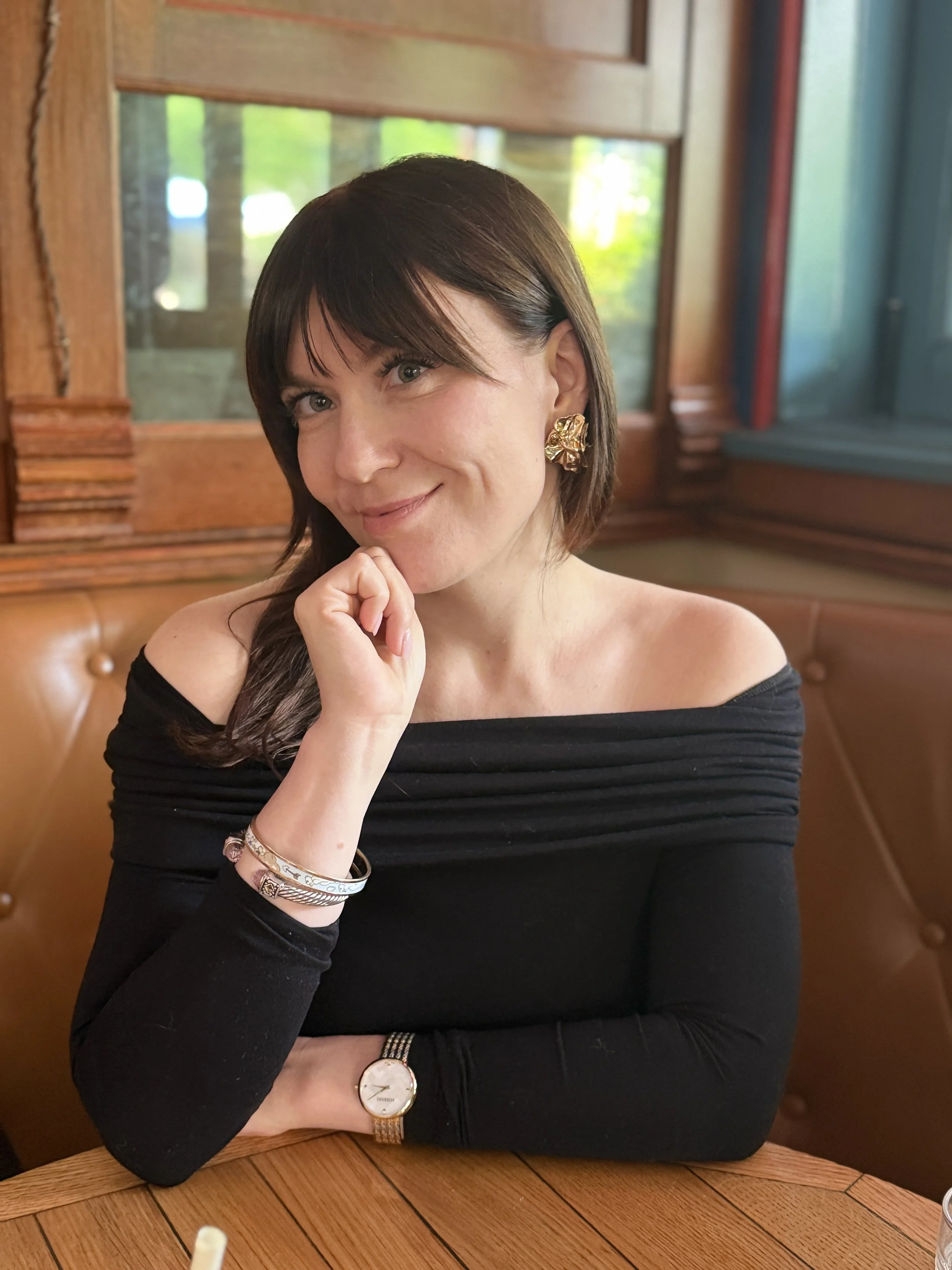 A woman with dark brown hair, wearing a black off-the-shoulder top, large gold earrings, a gold watch, and several bracelets, sitting at a wooden table in a restaurant or cafe with wood-paneled walls and a window with greenery outside.