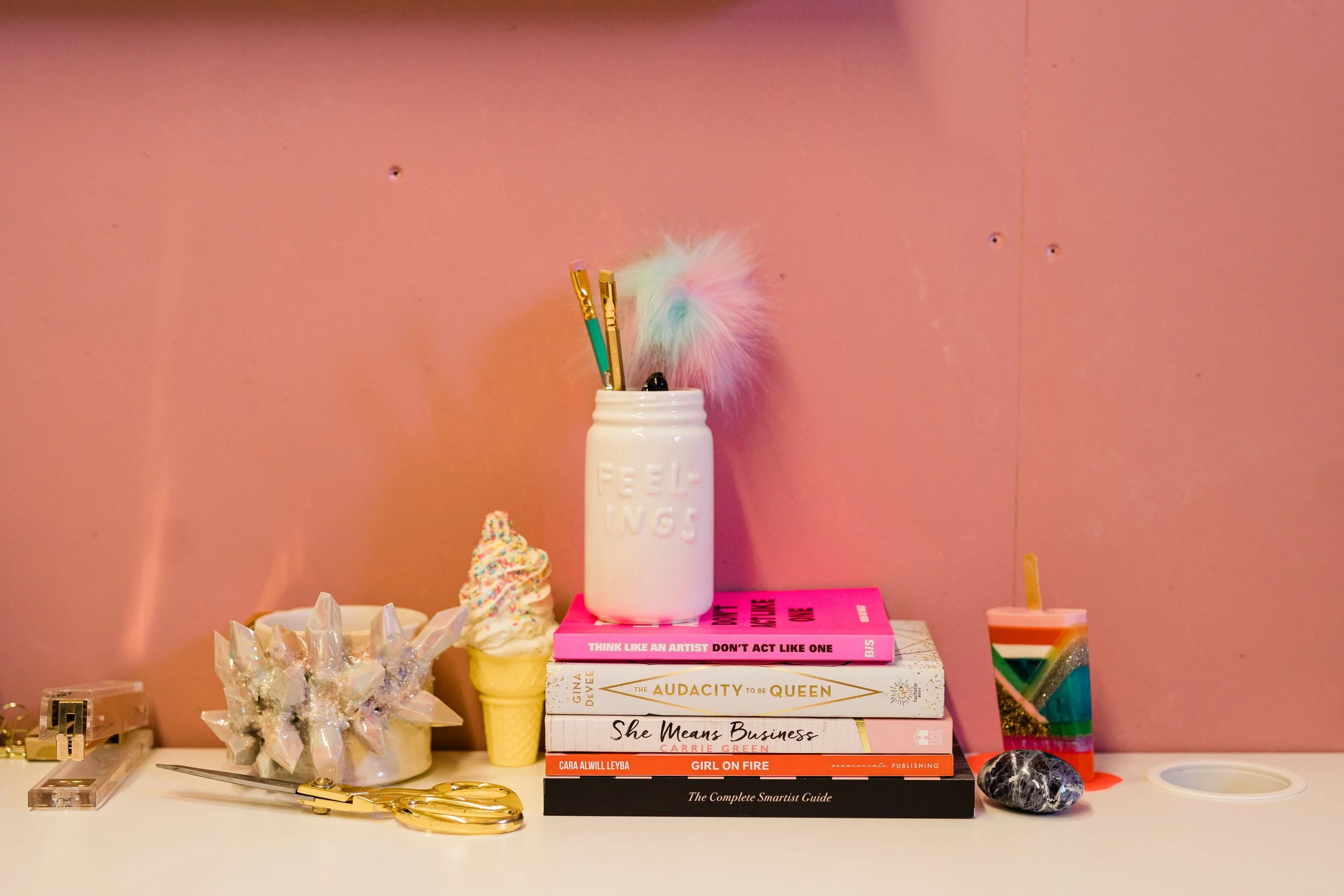 Decorative desk with pink wall background, including a white jar labeled 'FEELINGS' holding pastel-colored pens and a fluffy pom-pom, a stack of books about art, leadership, and business, a rainbow-colored candle, a black and white stone, a pair of gold scissors, a small decorative box, and a colorful crystal-shaped object.