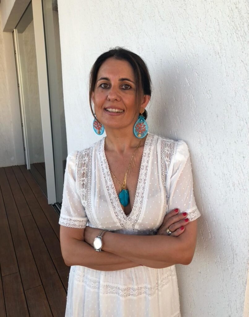A woman with dark hair, wearing a white lace dress, colorful earrings, a blue pendant necklace, a silver watch, and rings, standing against a white textured wall on a wooden deck.