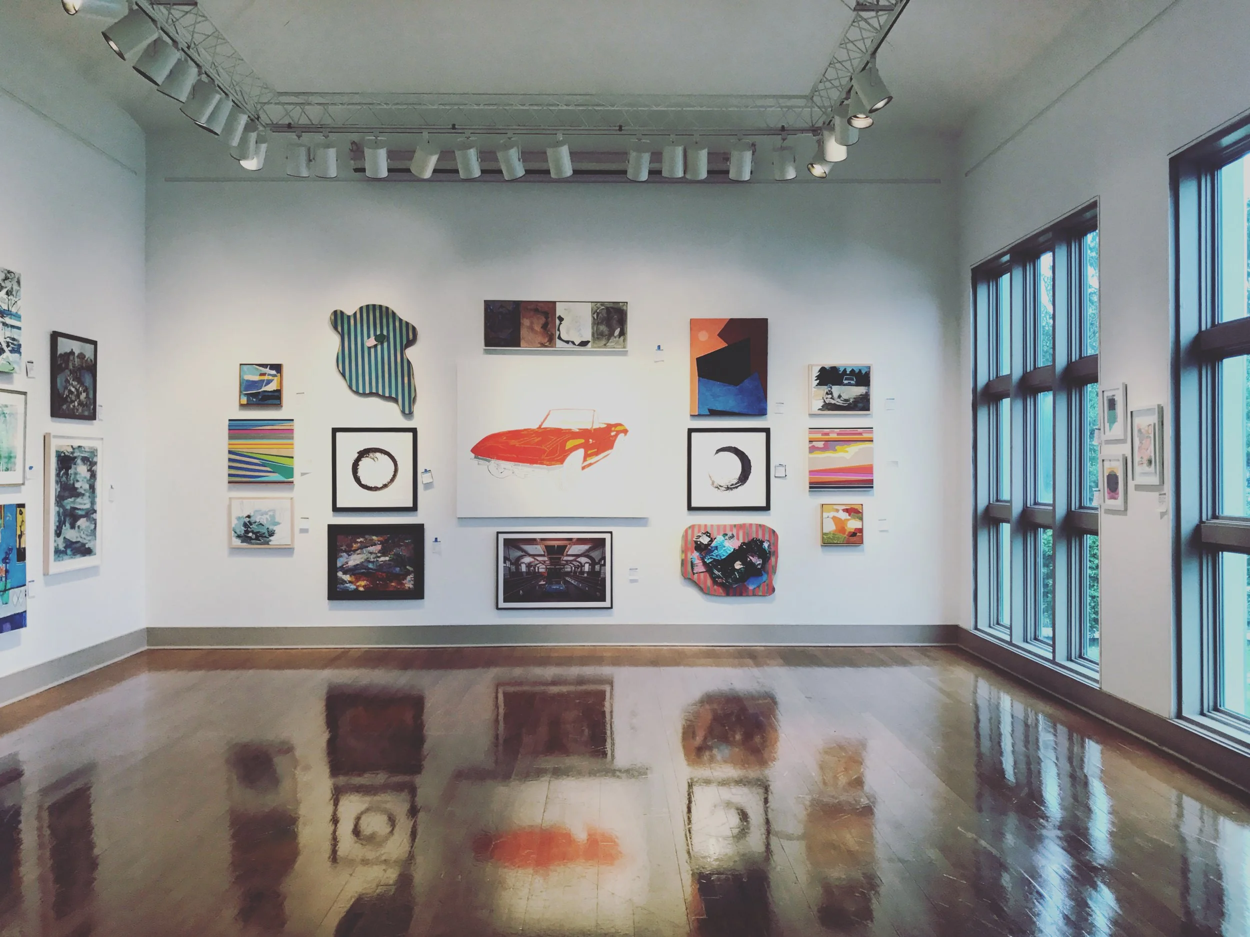 An art gallery with white walls and hardwood floors showcasing various colorful paintings and artworks, with large windows on the right side letting in natural light.