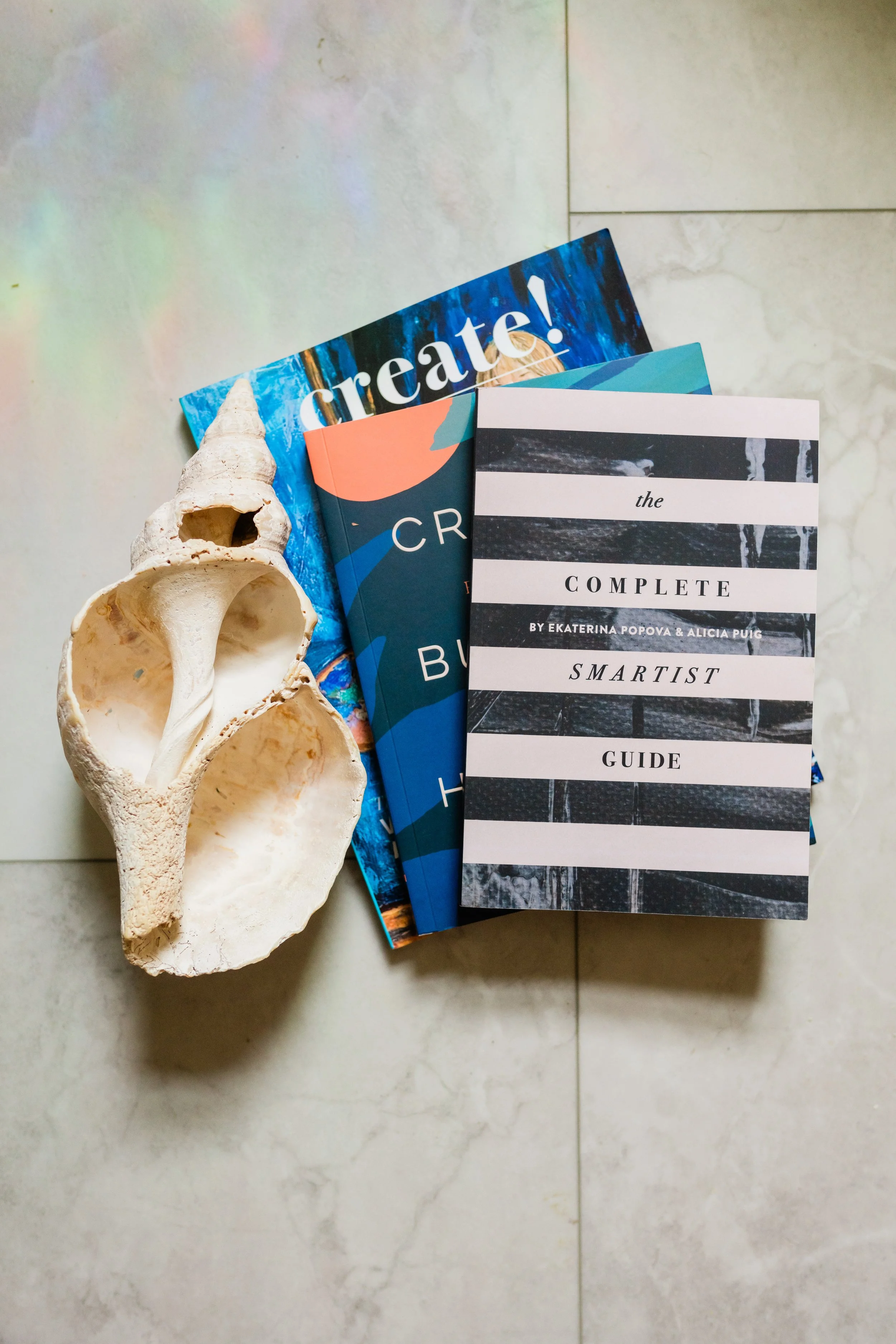 A seashell with a large spiral opening, overlapping books titled 'Creative' and 'The Complete Smartist Guide', placed on a light-colored tiled floor.