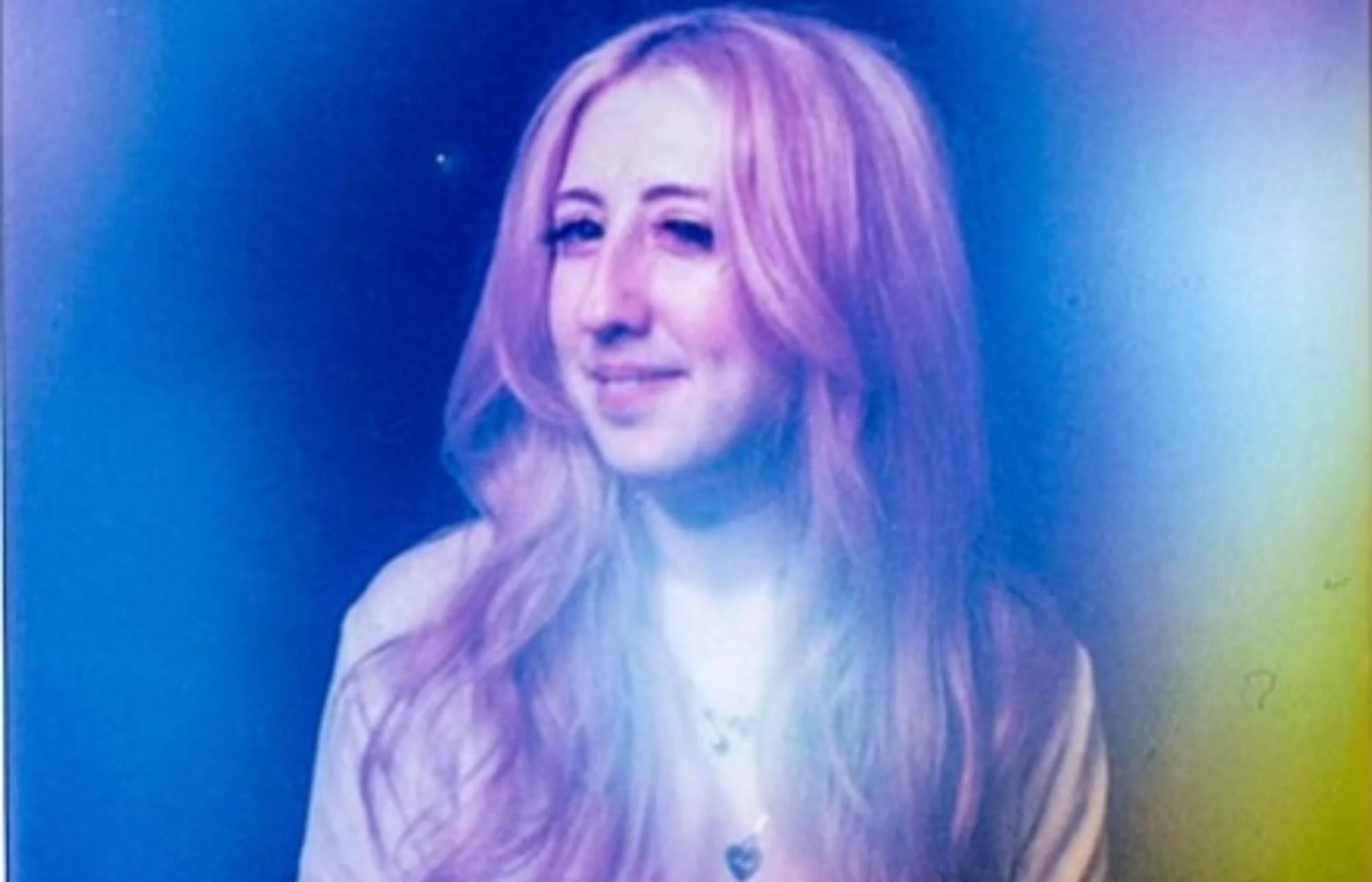 A woman with long, wavy, pinkish-purple hair smiling and winking in a studio with a dark background.