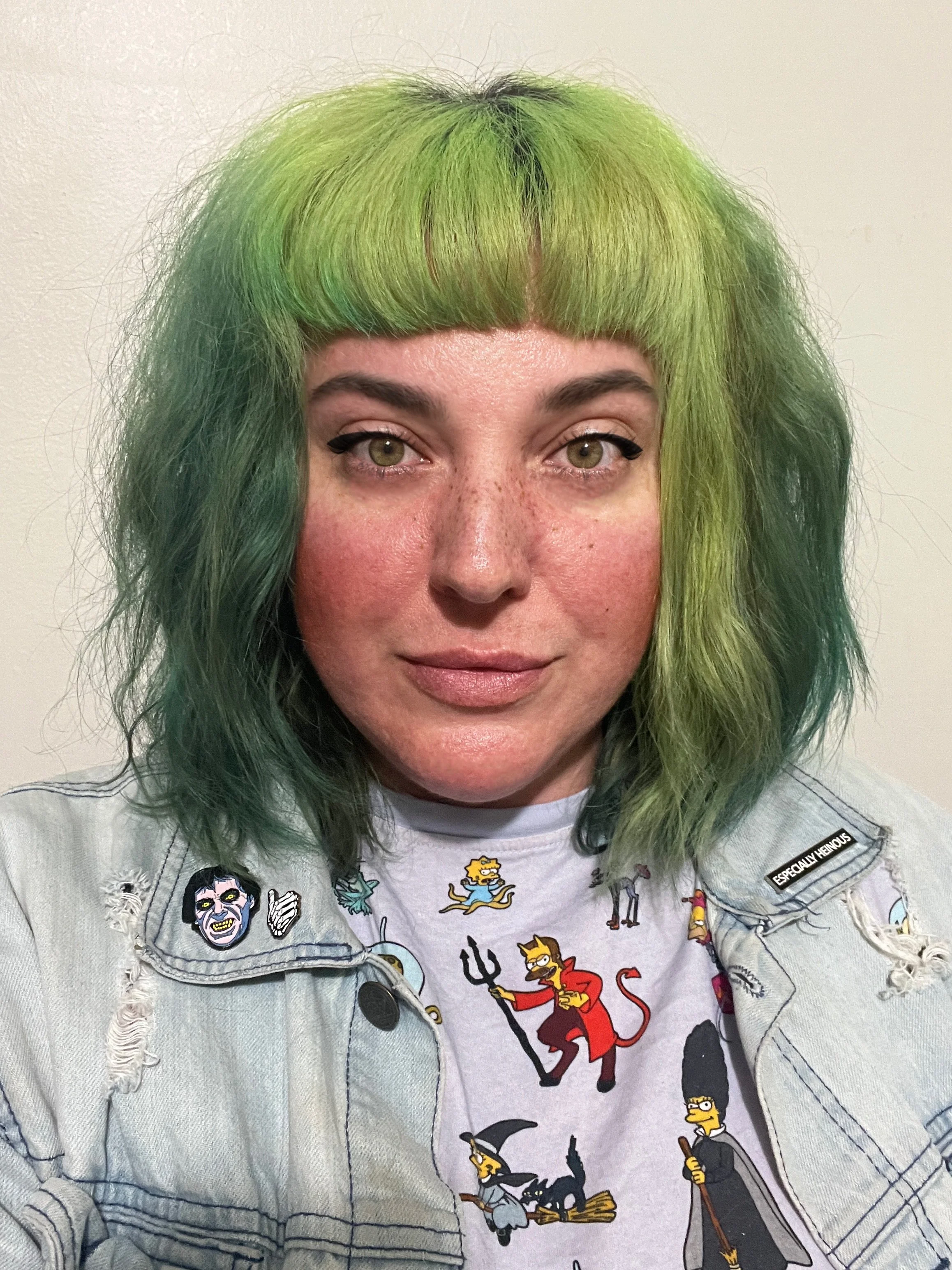 A person with green hair and light skin wearing a light denim jacket and a white shirt with cartoon character prints. The person has freckles and is looking directly at the camera.
