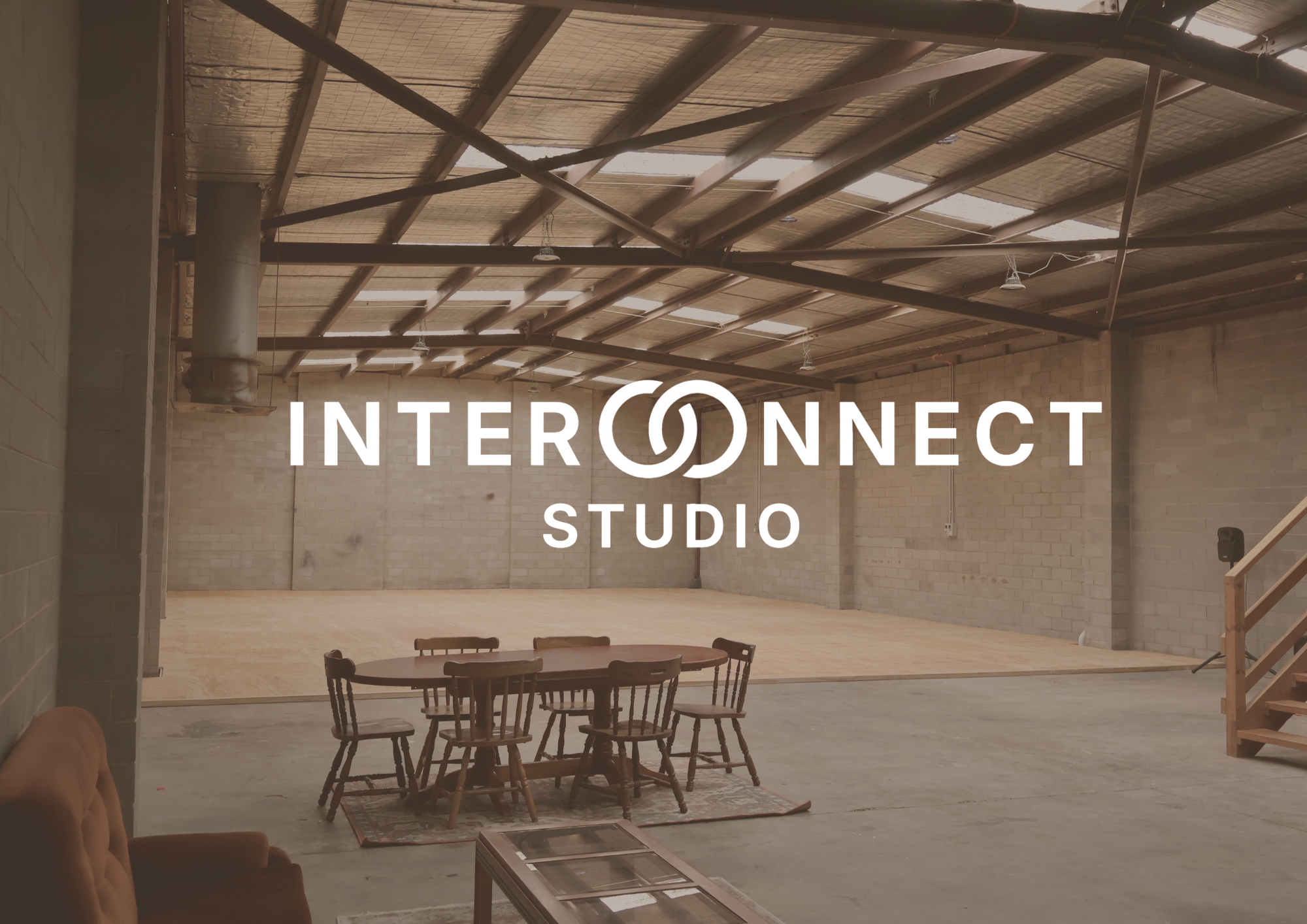 An empty studio with a wooden floor and a high ceiling made of metal beams and panels. There are a few tables and chairs in the space, with some furniture partially visible. The words 'INTERCONNECT STUDIO' are overlaid on the image.