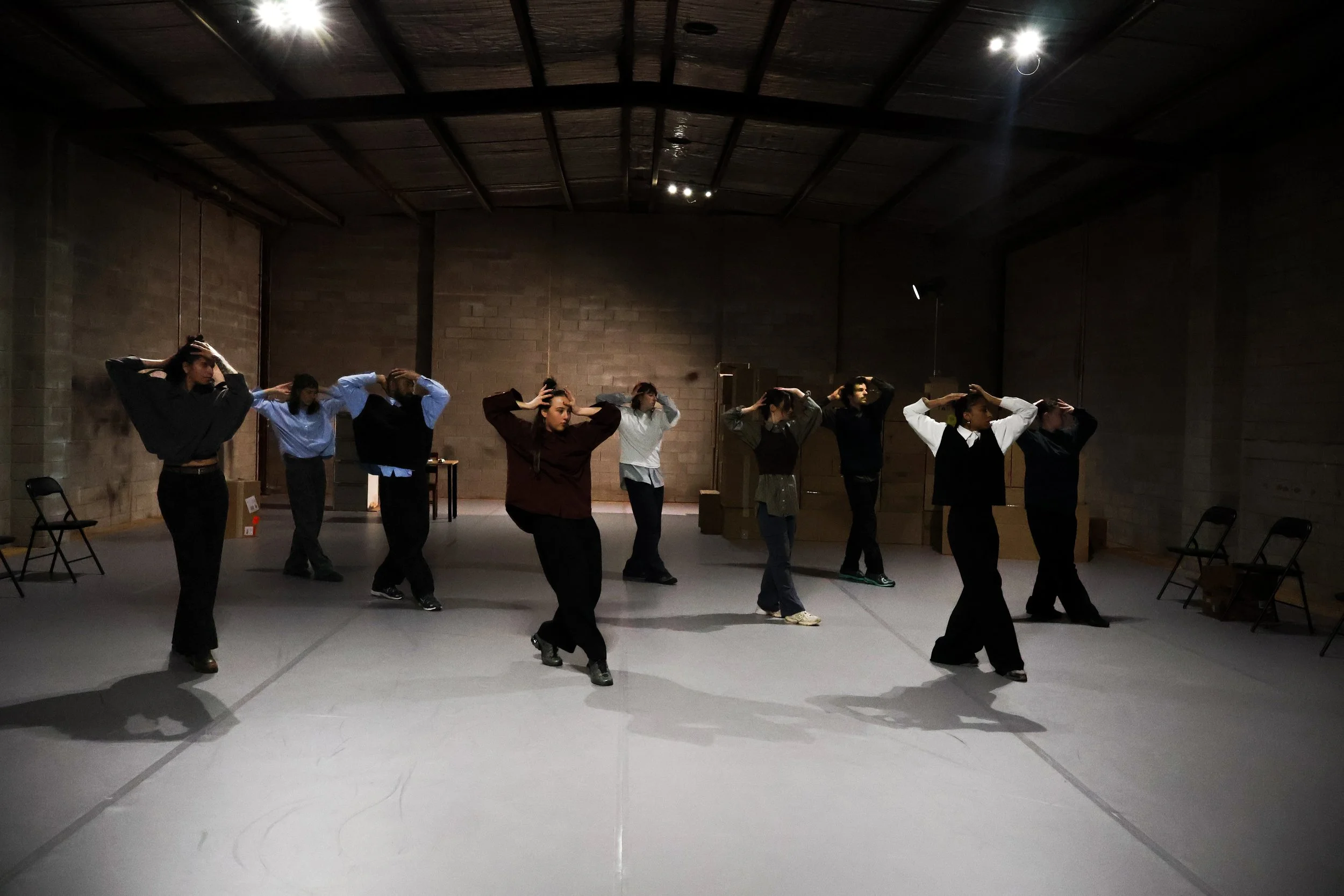 Group of people practicing dance or theatrical performance in a dimly lit studio with brick walls, some with hands on their heads, facing different directions.
