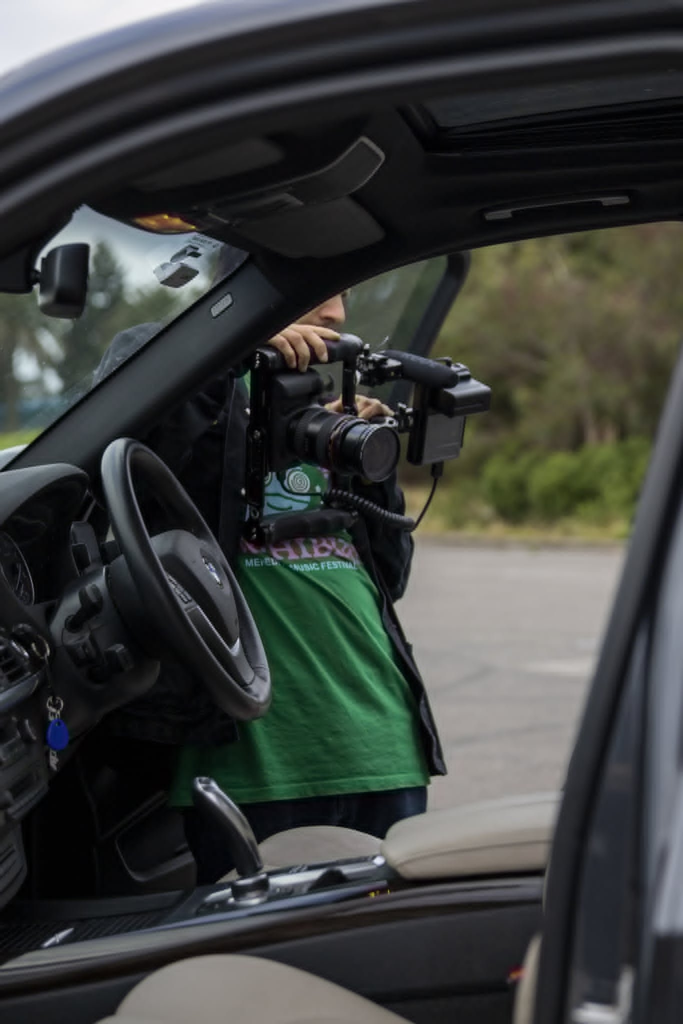 A person wearing a green shirt and a dark jacket is seen through the open window of a vehicle, holding a professional camera with a large lens, and appears to be taking pictures of something outside.