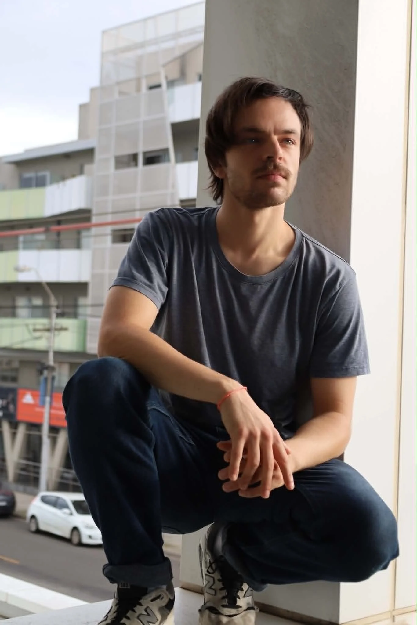 A young man with brown hair and a beard sitting on a window ledge with a city view in the background. He is wearing a dark t-shirt, dark pants, and Whitestone sneakers. The photo is taken during daytime.