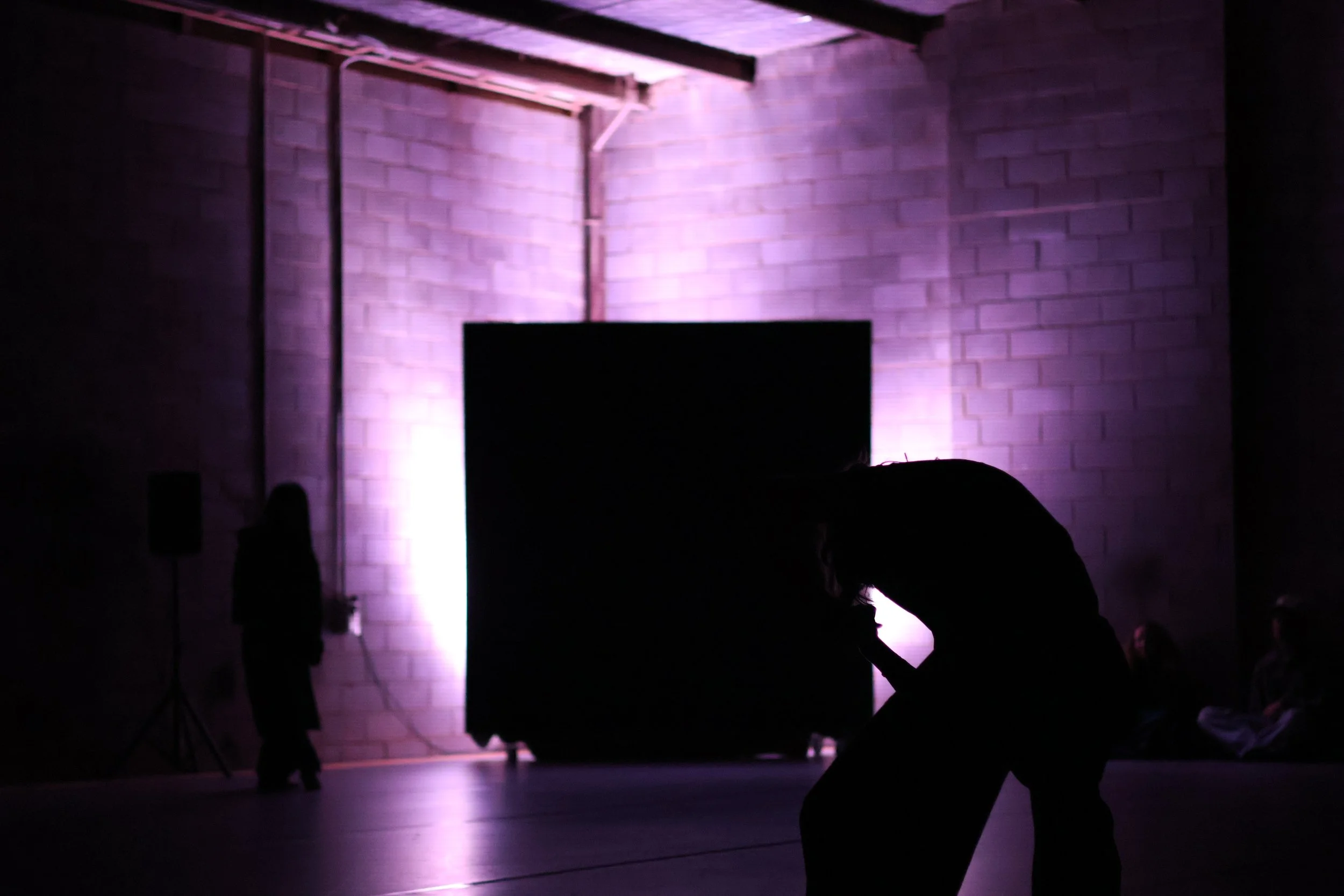 A dark room with a stage illuminated by purple lights. Several silhouettes of people are present, some facing the stage and others engaged in different activities.