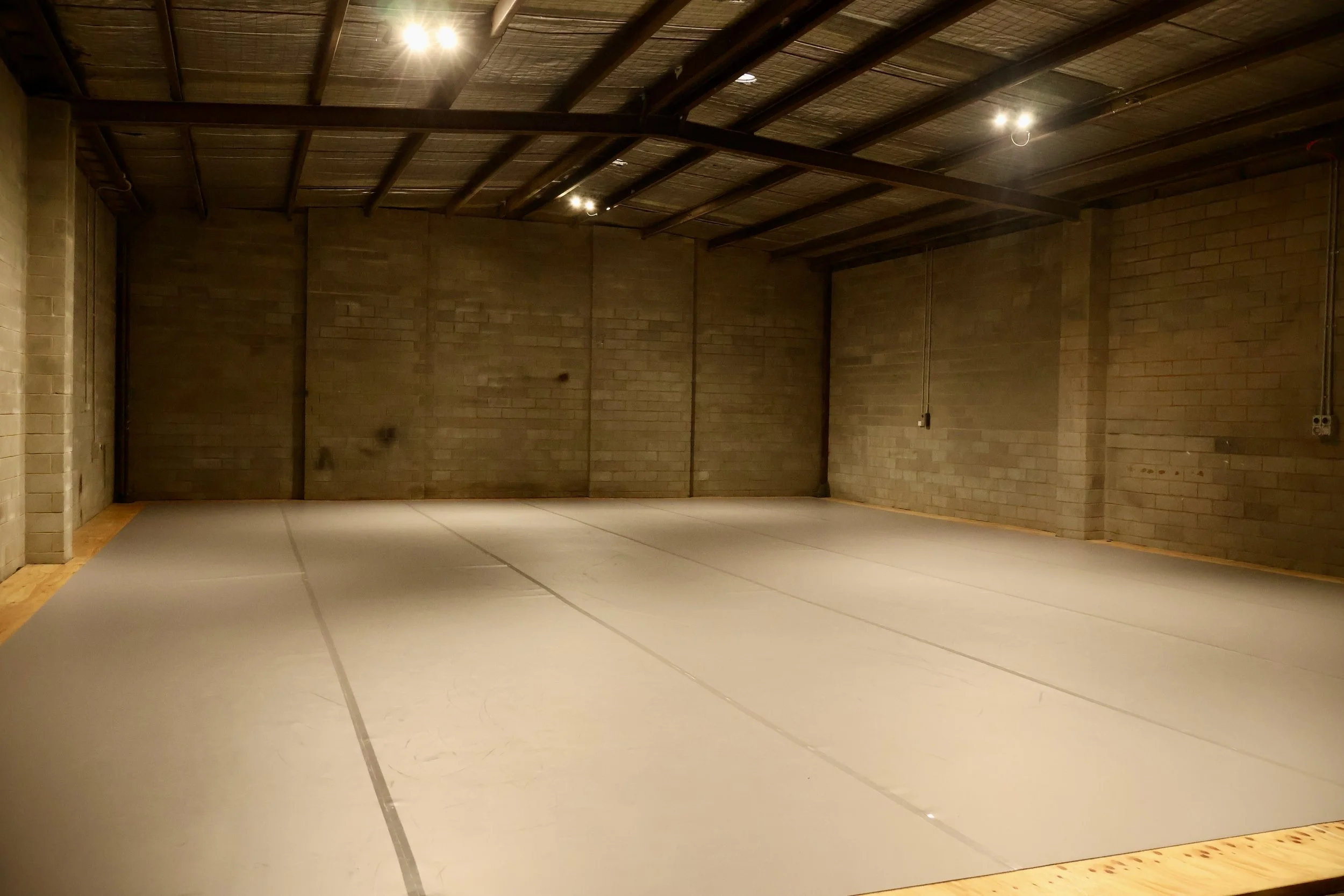 An empty indoor dojo with beige brick walls, a wooden floor border, and a concrete floor with gray mats. The ceiling has exposed wooden beams and lights.