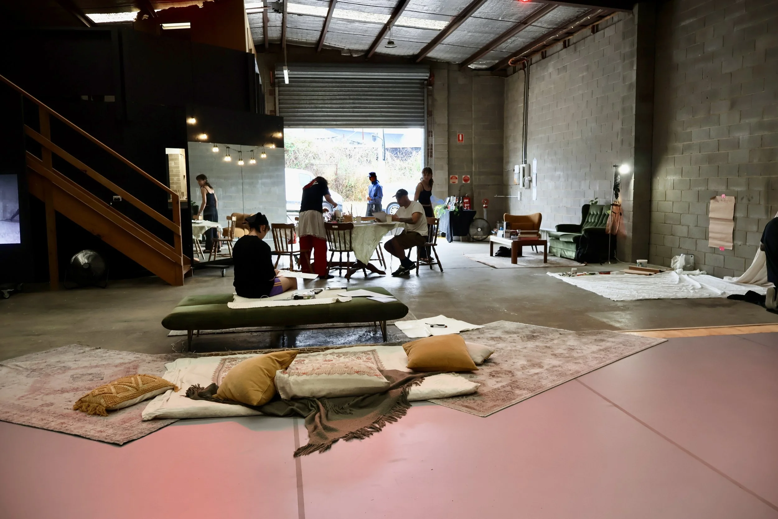 People in an industrial-style indoor space with a high ceiling, working around tables and on the floor. There are pillows and rugs on the floor, and the space appears to be set up for an art project or workshop.