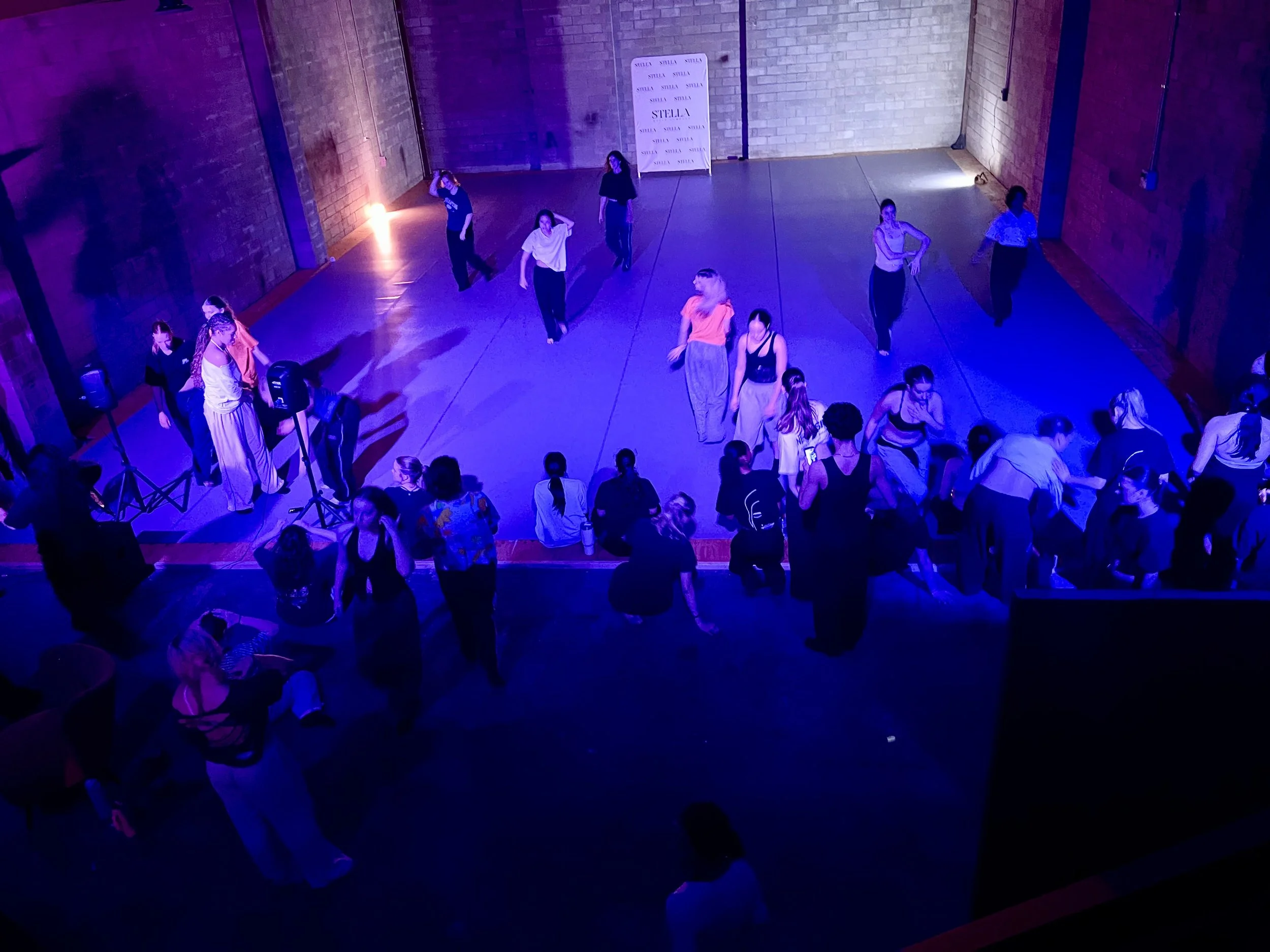 Dance studio with a group of people dancing under purple and blue stage lights, with brick walls and a large white banner in the background.