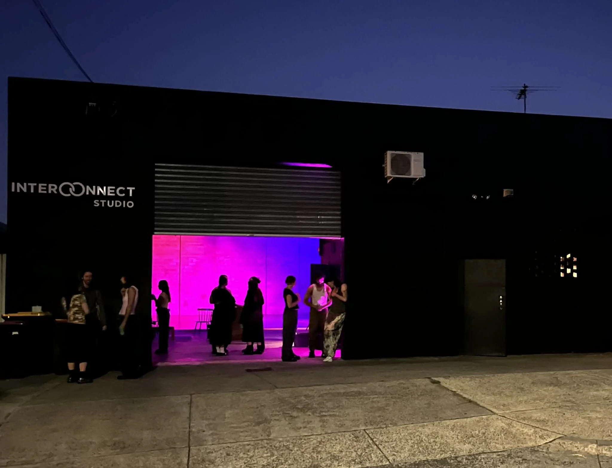 People gathered outside a building at night, with neon pink and purple lighting illuminating the entrance, and a sign reading 'INTERCONNECT STUDIO' on the side of the building.