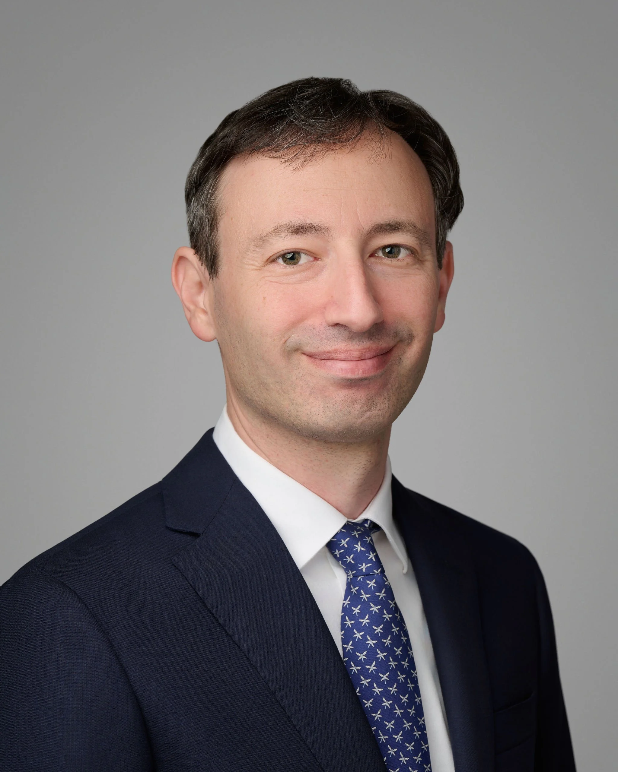 A professional headshot of a man in a dark suit with a white shirt and a blue tie with white star patterns, smiling slightly against a gray background.