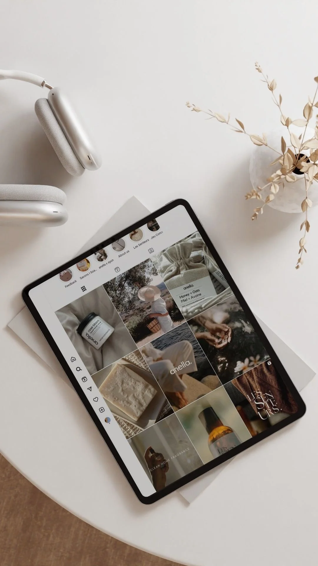 An iPad displaying a social media grid of skincare and lifestyle images on a white desk, with white headphones and a white vase with dried leaves nearby.