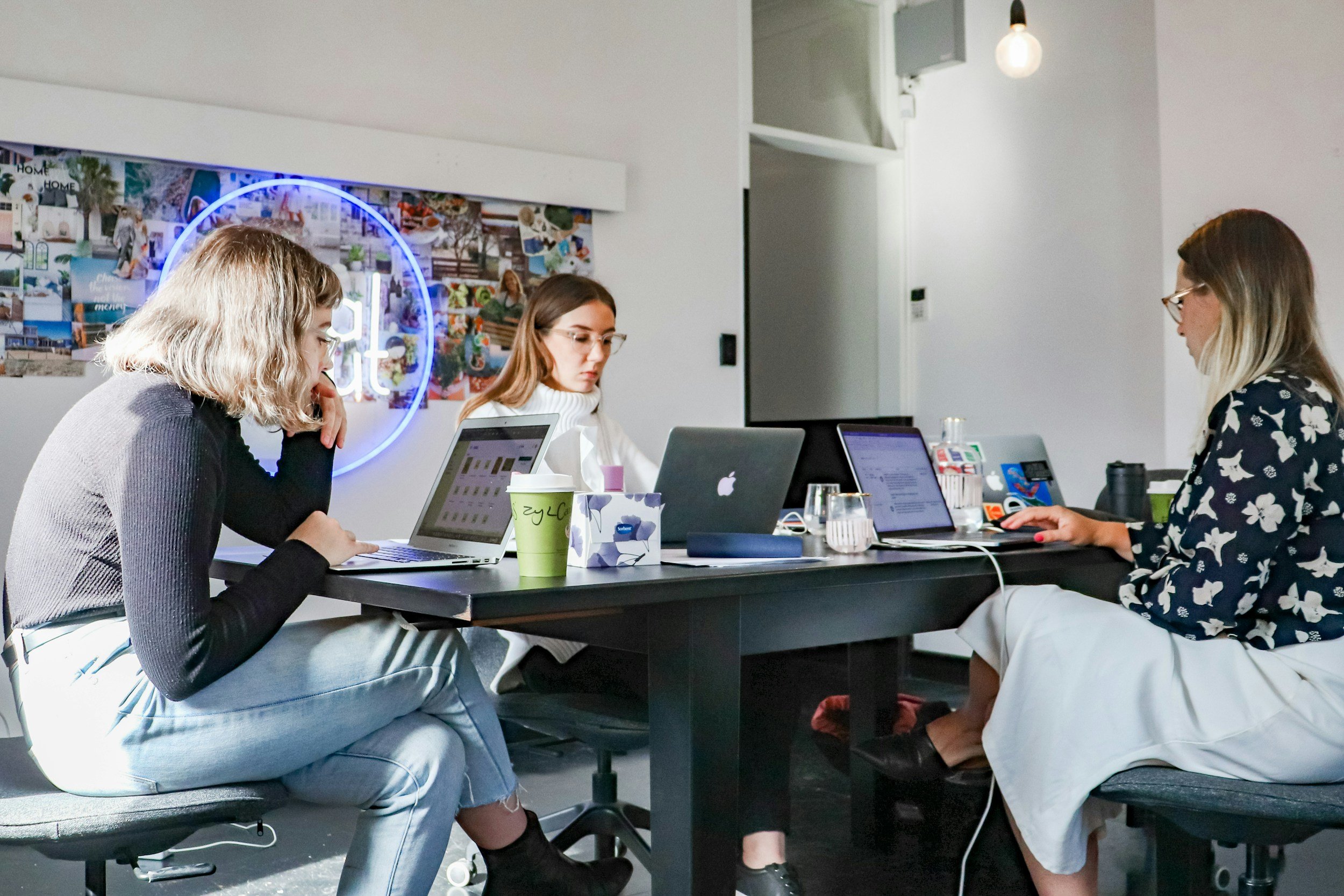 Three women working on laptops at a conference table in an office with a colorful collage and neon sign in the background.