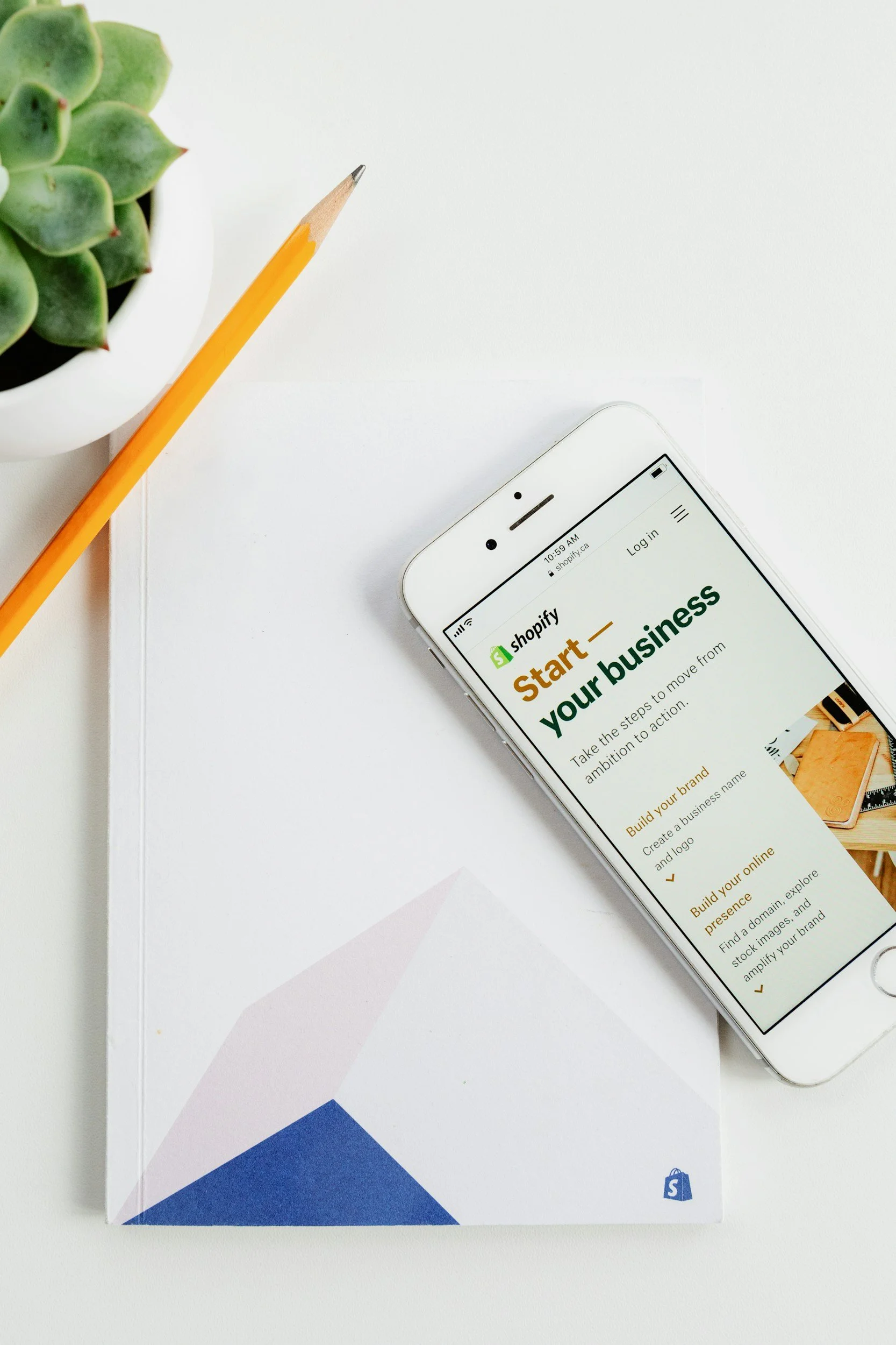 Smartphone displaying Shopify startup webpage, on a desk with a yellow pencil, succulent plant in a white pot, and a white paper with blue and purple geometric sections.