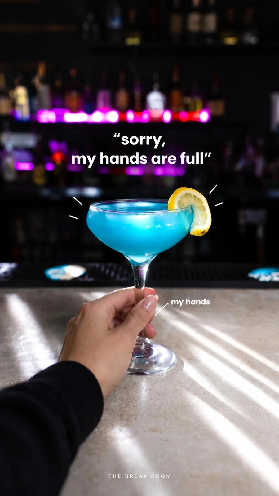 A hand holding a blue cocktail with a lemon wedge garnish on the rim, set on a bar counter. The background is blurred with neon lights and bottles. The image features a quote: 'sorry, my hands are full' and labels pointing to 'my hands' and 'the bar'