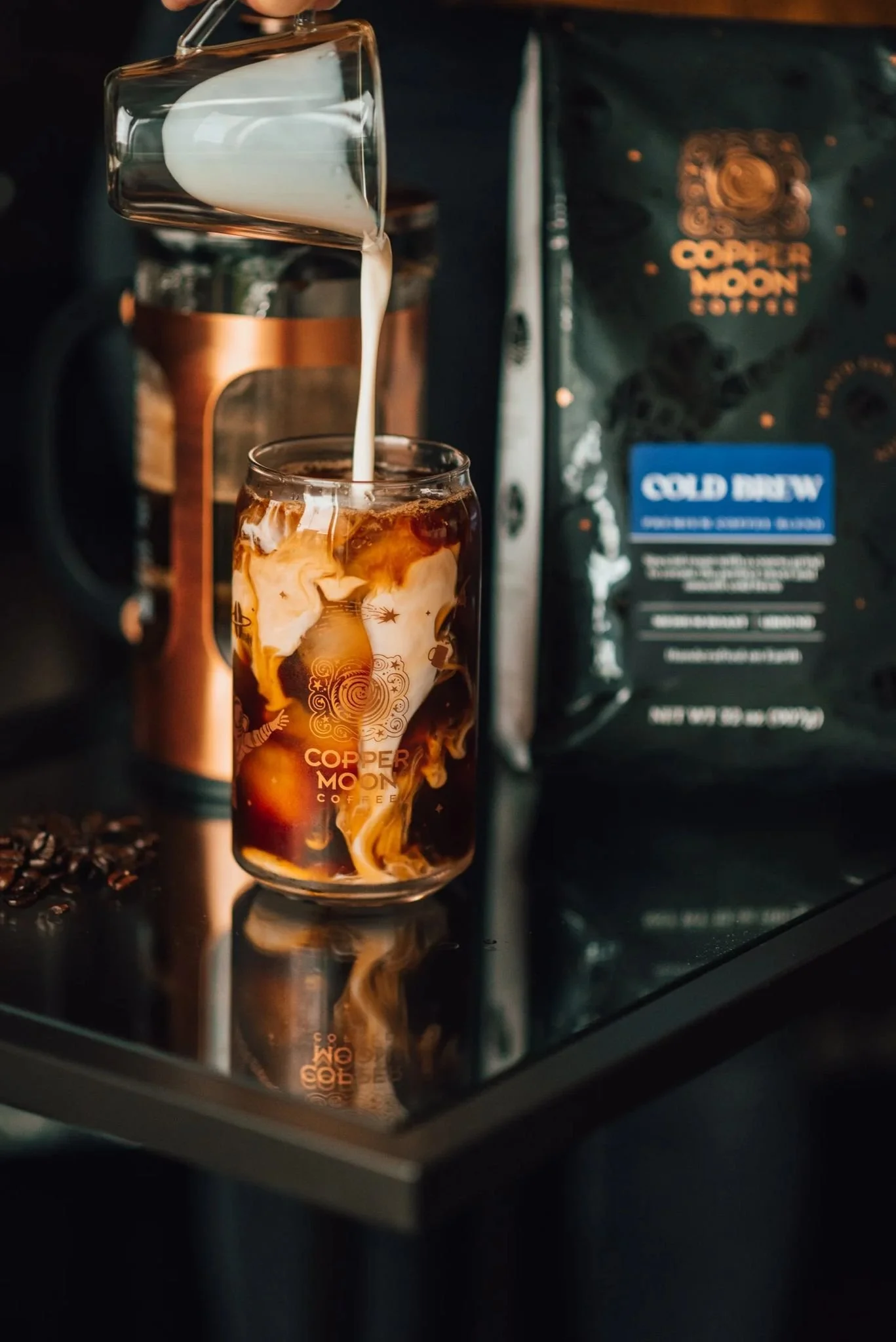 A glass of iced coffee with cream swirling in it, on a reflective surface, with a hand pouring cream from a small glass pitcher. In the background, there is a bag of Copper Moon Coffee labeled 'Cold Brew' and a copper-colored coffee maker.