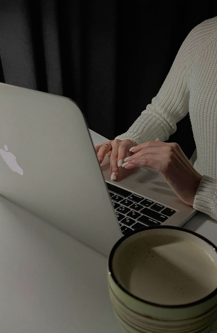 Close-up of a person typing on a silver MacBook laptop at a white table, with a partially visible coffee mug in the foreground.