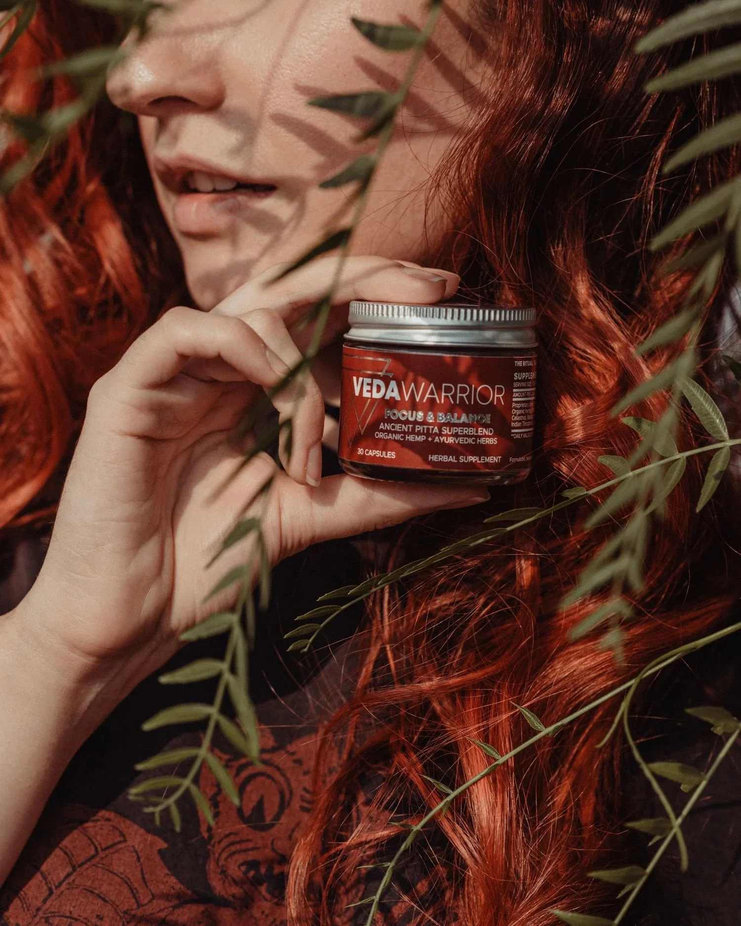 A woman with long, curly red hair holds a container labeled 'Veda Warrior Focus & Balance' herbal supplement, surrounded by greenery.