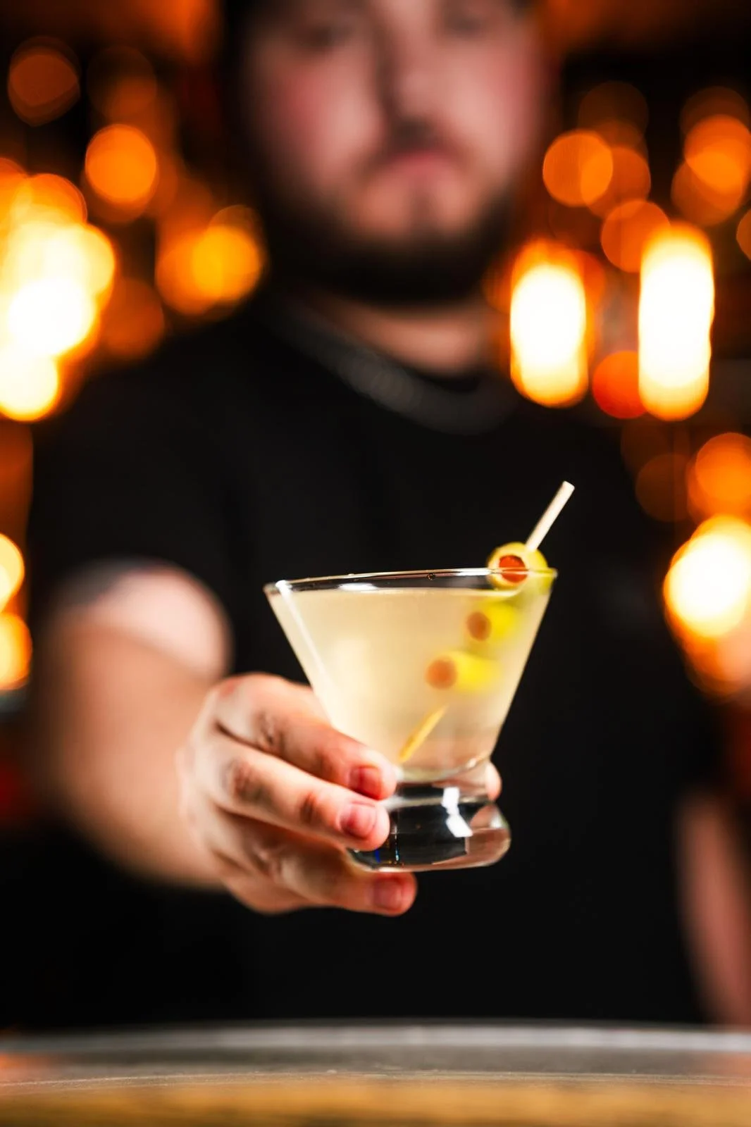 A man holding a martini glass with an olive garnish, blurred background with warm bokeh lights.