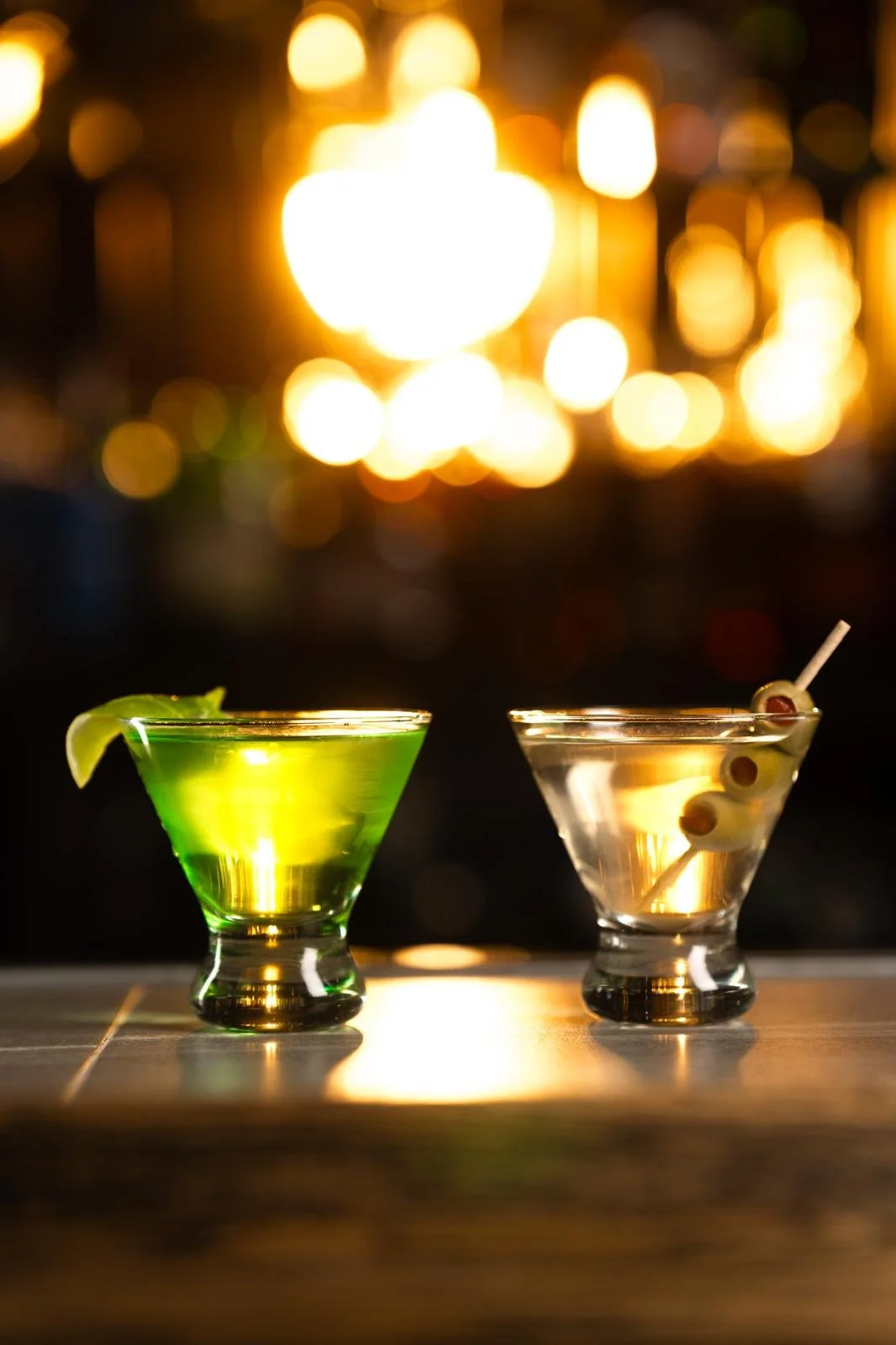 Two cocktails on a table, one green and one clear with olives, against a backdrop of blurred warm lights.