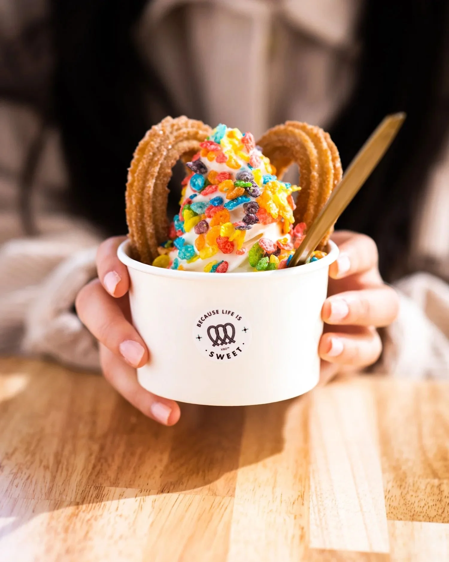A person holding a cup of soft serve ice cream topped with colorful cereal pieces and two churros, with a wooden spoon inside.