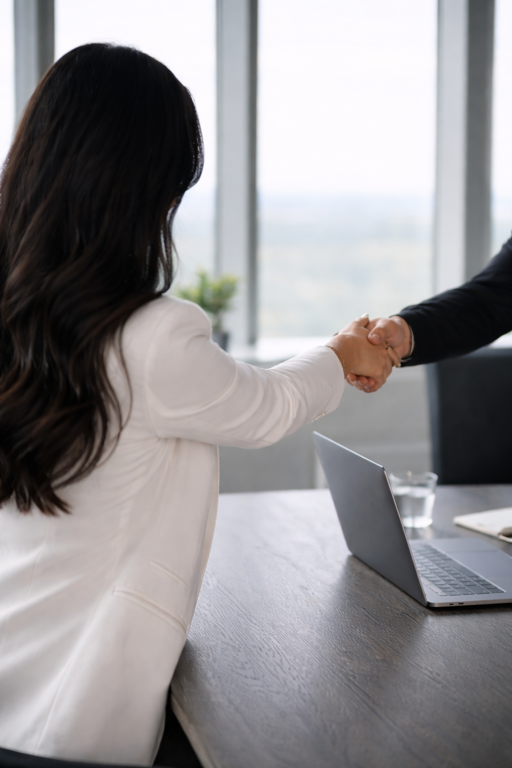 Woman in a white blazer shaking hands with a person in a black suit in a modern office setting.