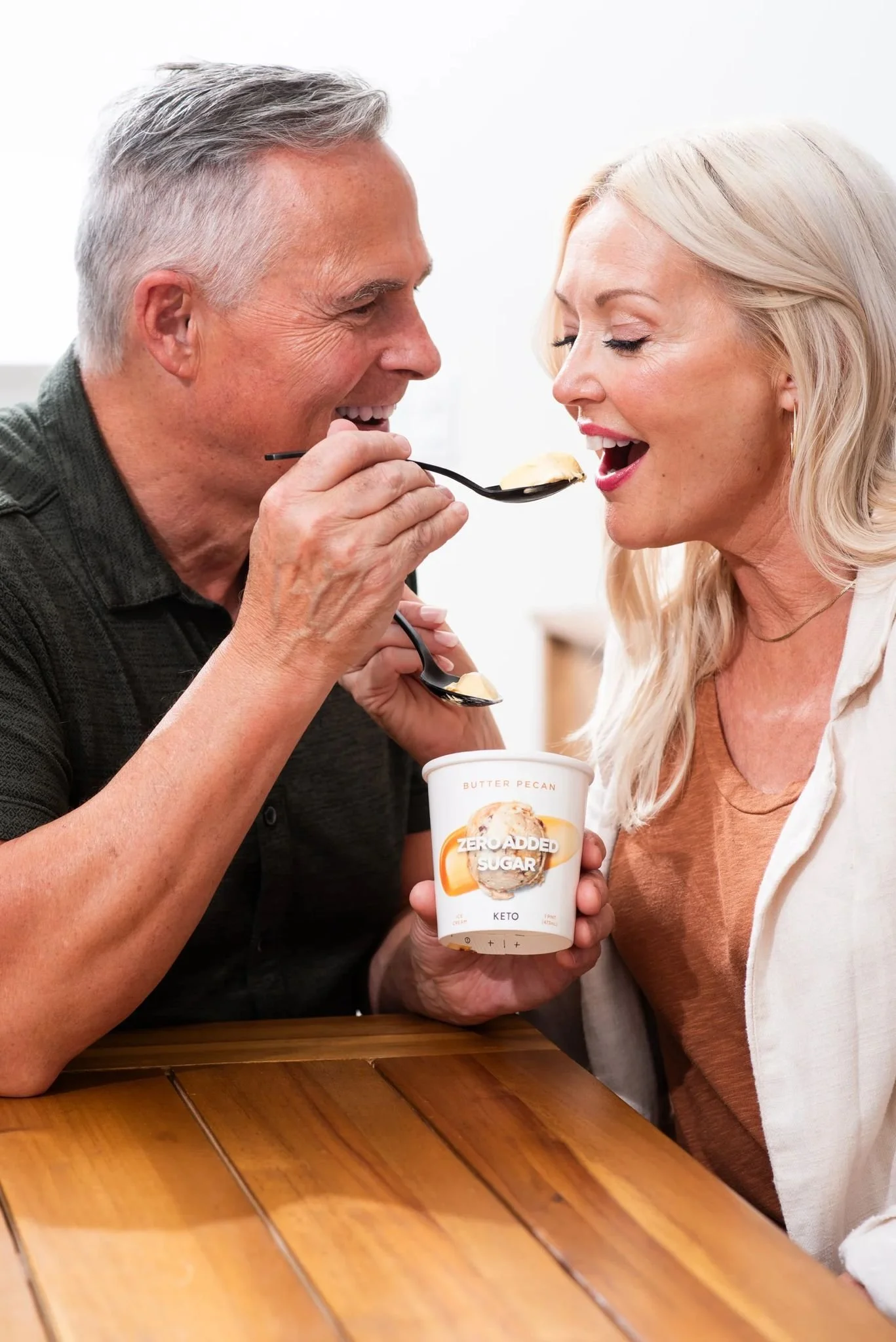 A man is feeding a woman with a spoon. The woman is holding a container labeled 'Zero Added Sugar' and 'Keto' with the flavor 'Butter Pecan.' They are sitting at a wooden table in a bright setting.